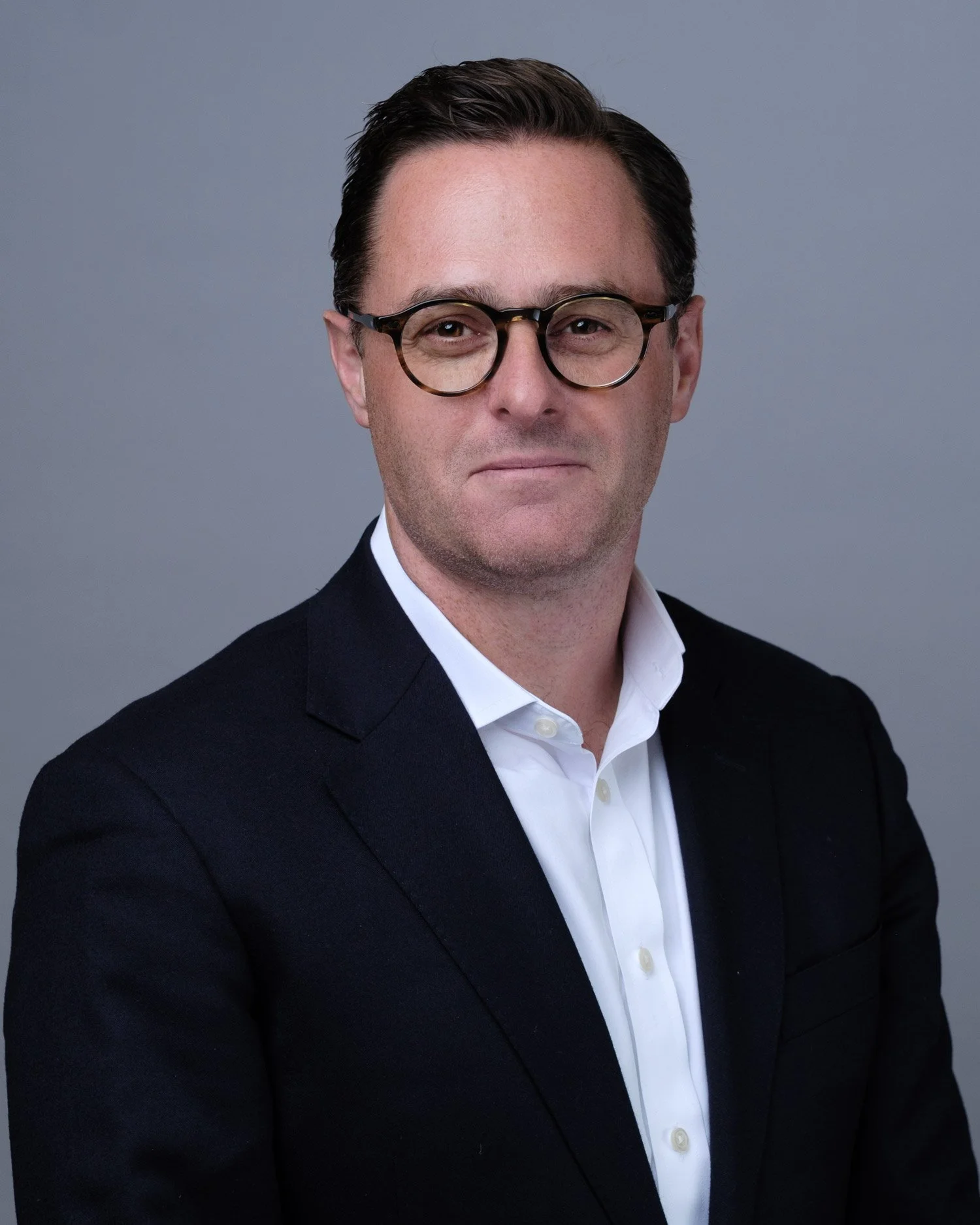 A man wearing a black suit, white shirt, and round glasses, posing against a plain gray background.