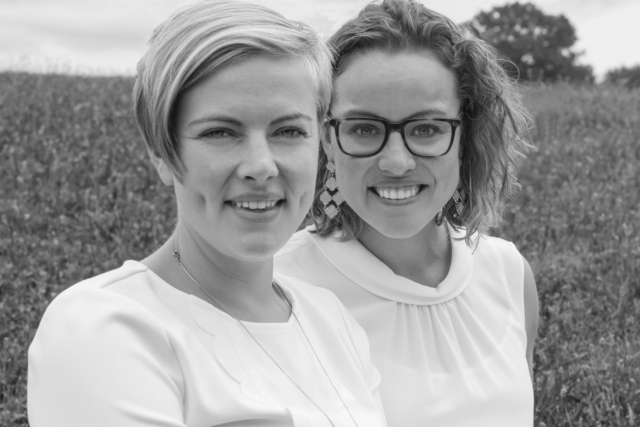 Two women standing close together outdoors, smiling at the camera, with a field and trees in the background.