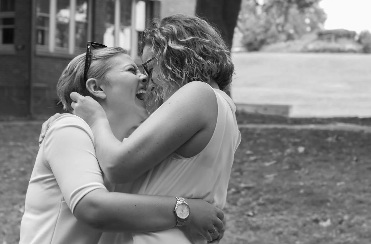 Two women are hugging and smiling happily outdoors, in a park or yard.