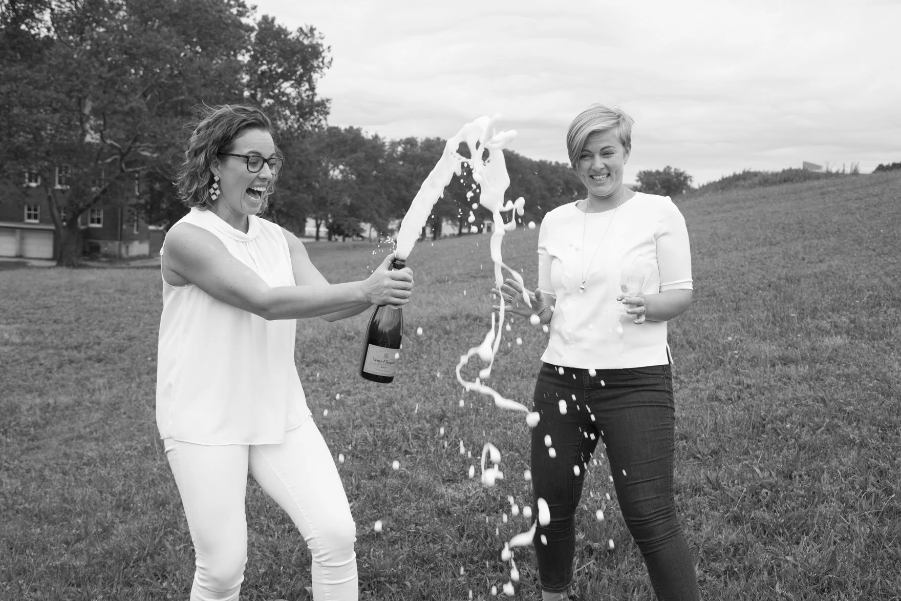 Two women celebrating outdoors with champagne, one opening the bottle with foam spraying out, both smiling and laughing on a grassy field with trees in the background.