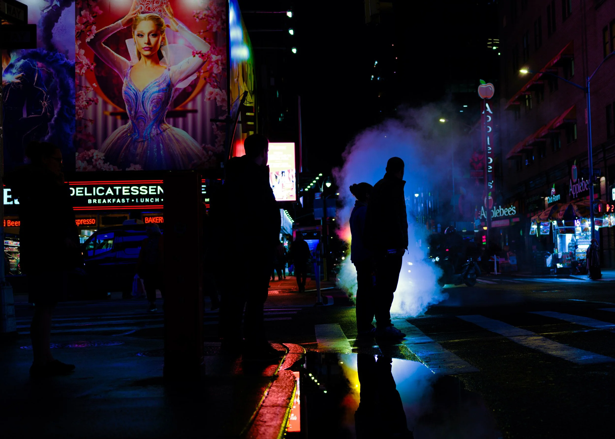 Nighttime street scene with illuminated billboards, people walking, and steam rising from the sidewalk. Visible advertisements include a large portrait of a woman and signs for a delicatessen and Applebee's restaurant.