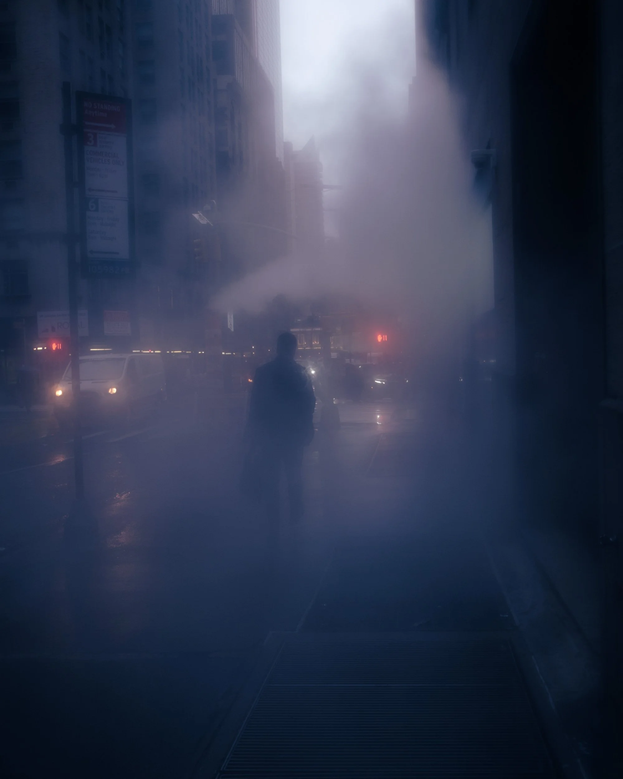 A city street enveloped in thick fog with silhouetted buildings and distant vehicles. The street has illuminated stop lights and signs, with a person walking in the foreground.