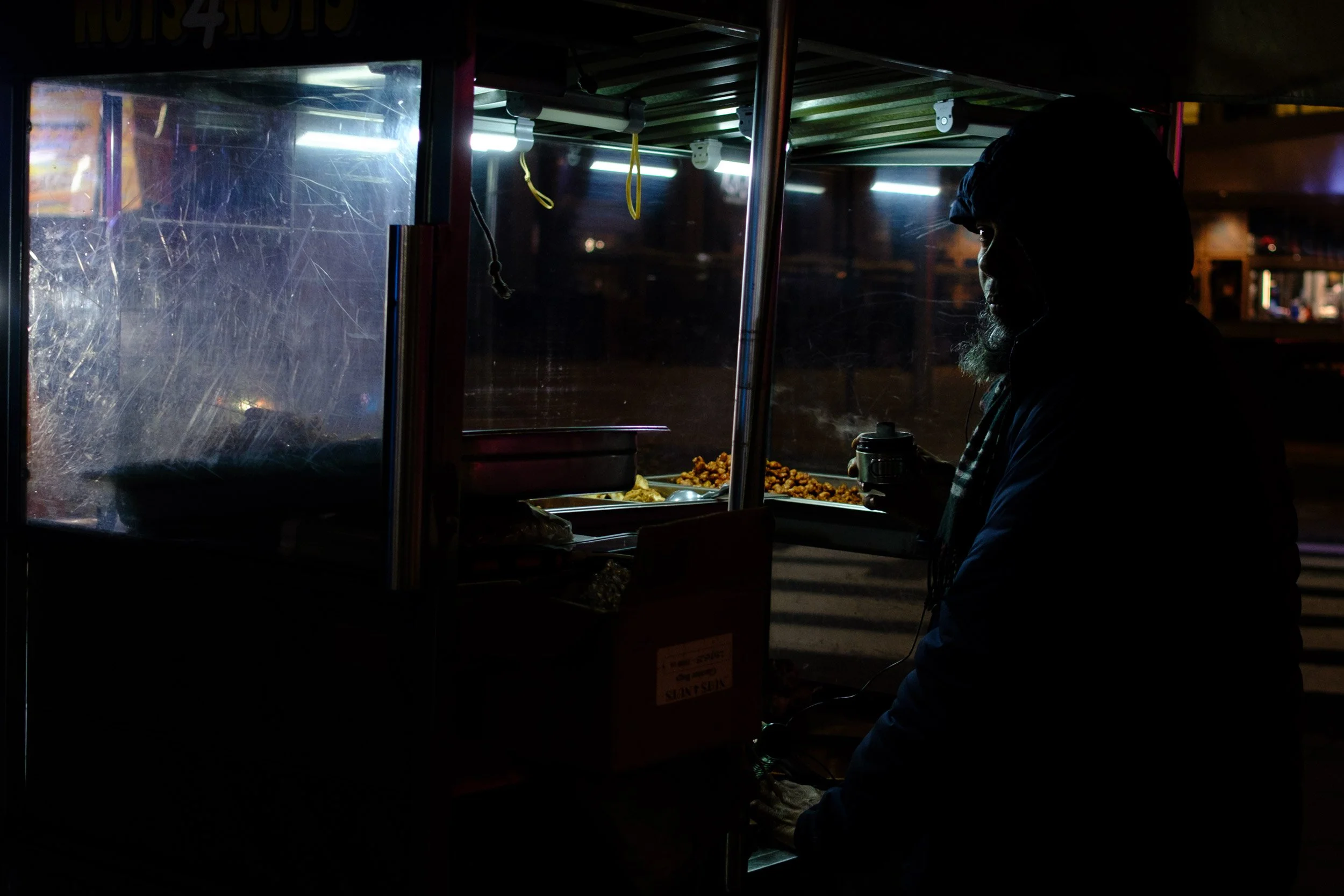 A person is selling food at a street food stall at night. The stall has a glass display case with some yellow hanging hooks and illuminated lighting inside, showing fried snacks. The person's face is partially visible in profile and they are wearing 
