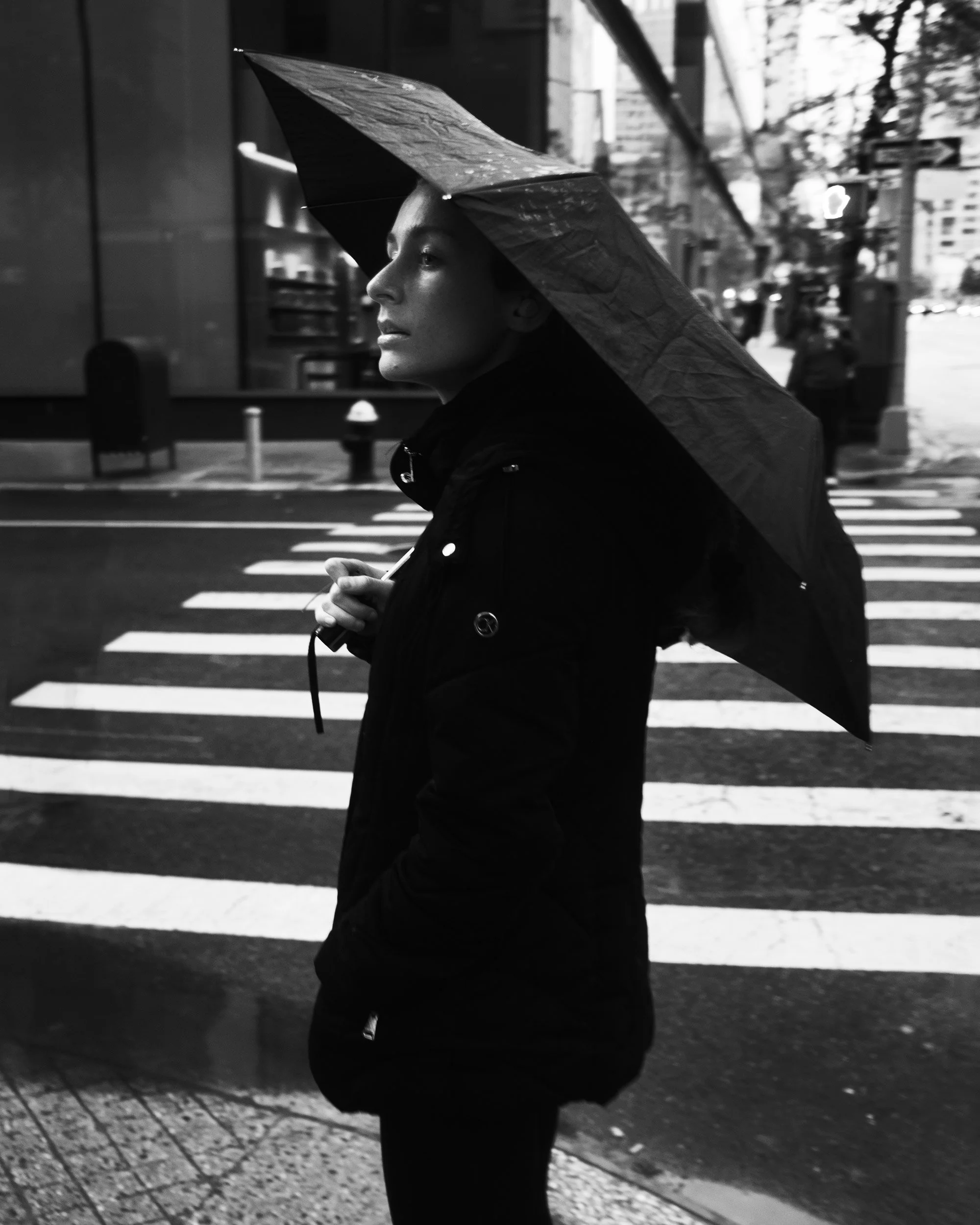 A woman wearing a jacket and holding an umbrella walks on a city street at night, with blurred background of buildings and trees.