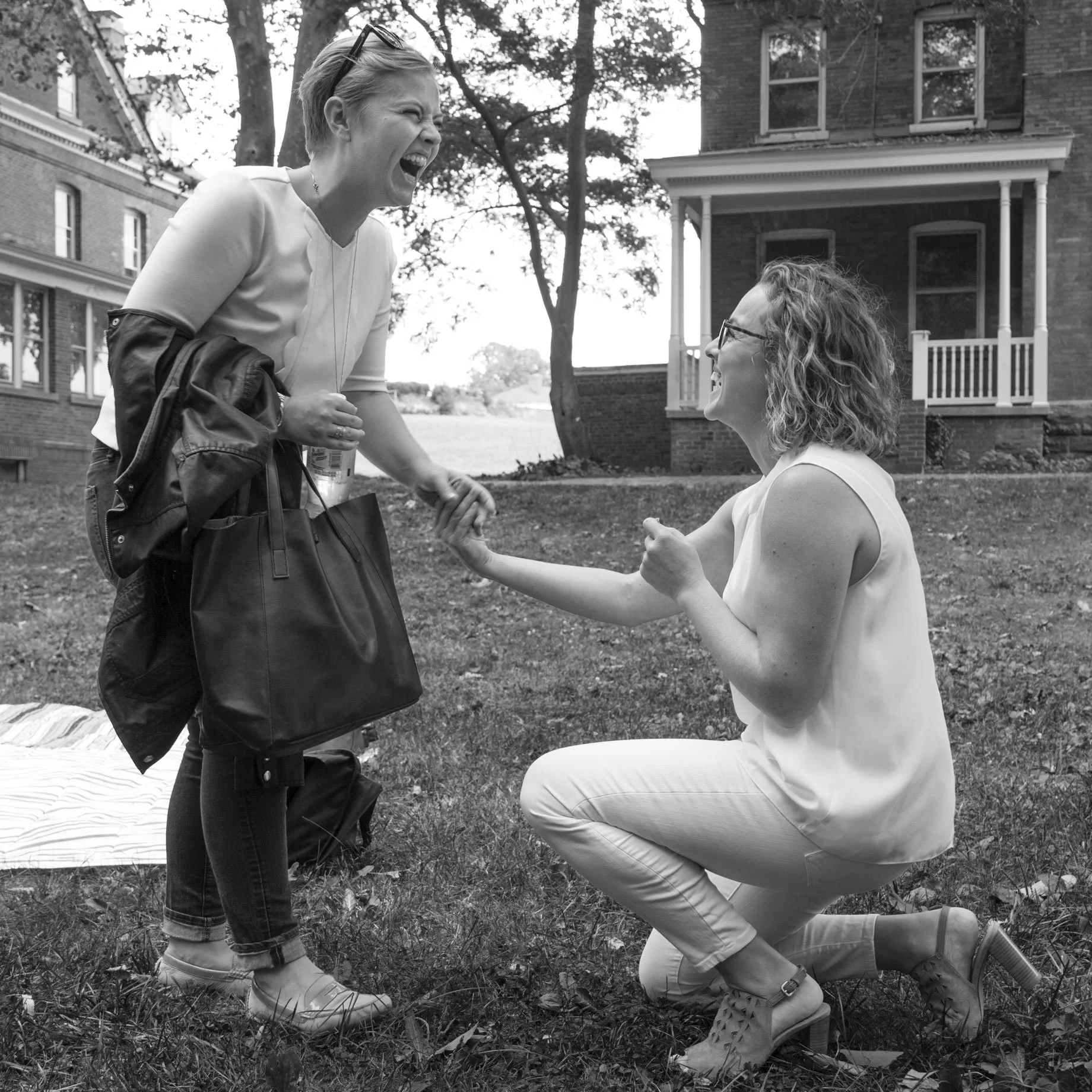 Two women are outdoors on a grassy area in front of brick houses, engaged in a joyful moment. One woman, standing, is laughing and holding a water bottle and purse, while the other woman, kneeling, is smiling and holding hands with the standing woman