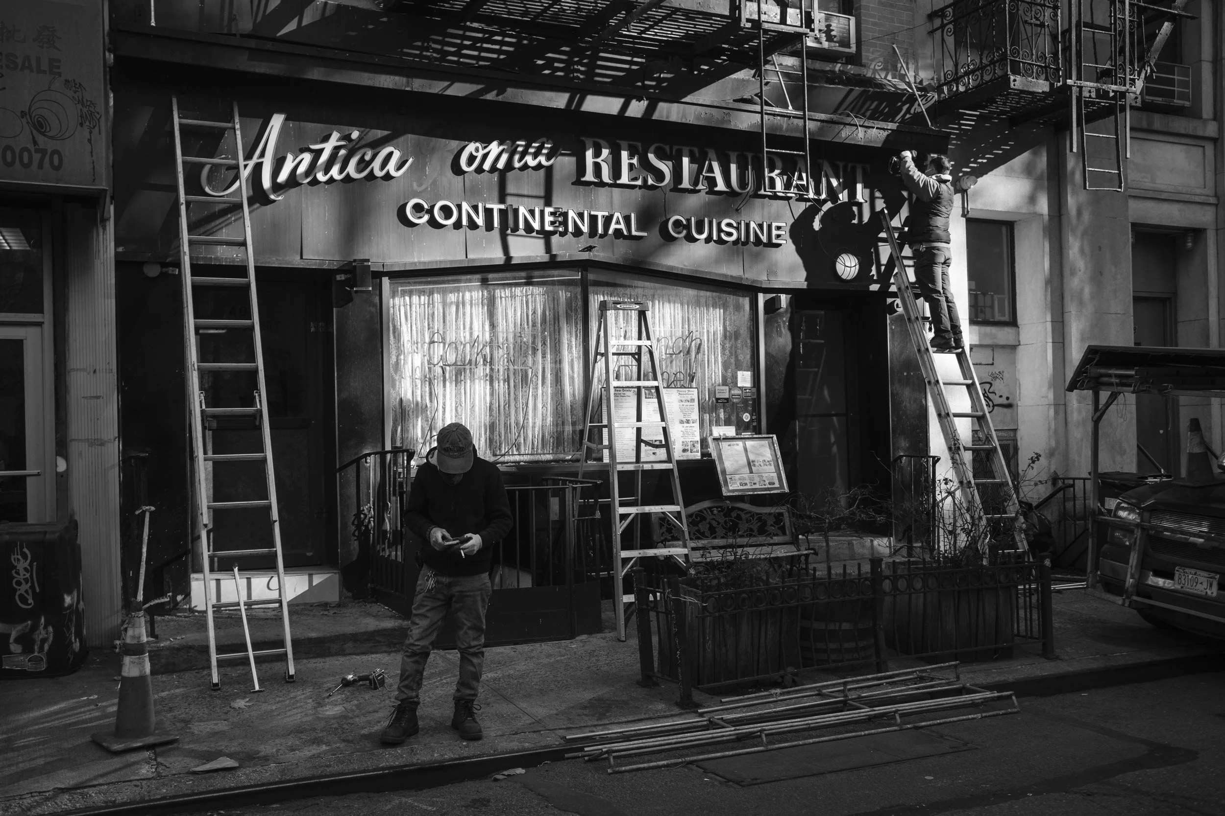 Two workers are installing or repairing a sign for an Italian restaurant called 'Antica Ora' on a city building. One worker is on a ladder, handling the sign, while the other is standing on the sidewalk, looking at his phone.