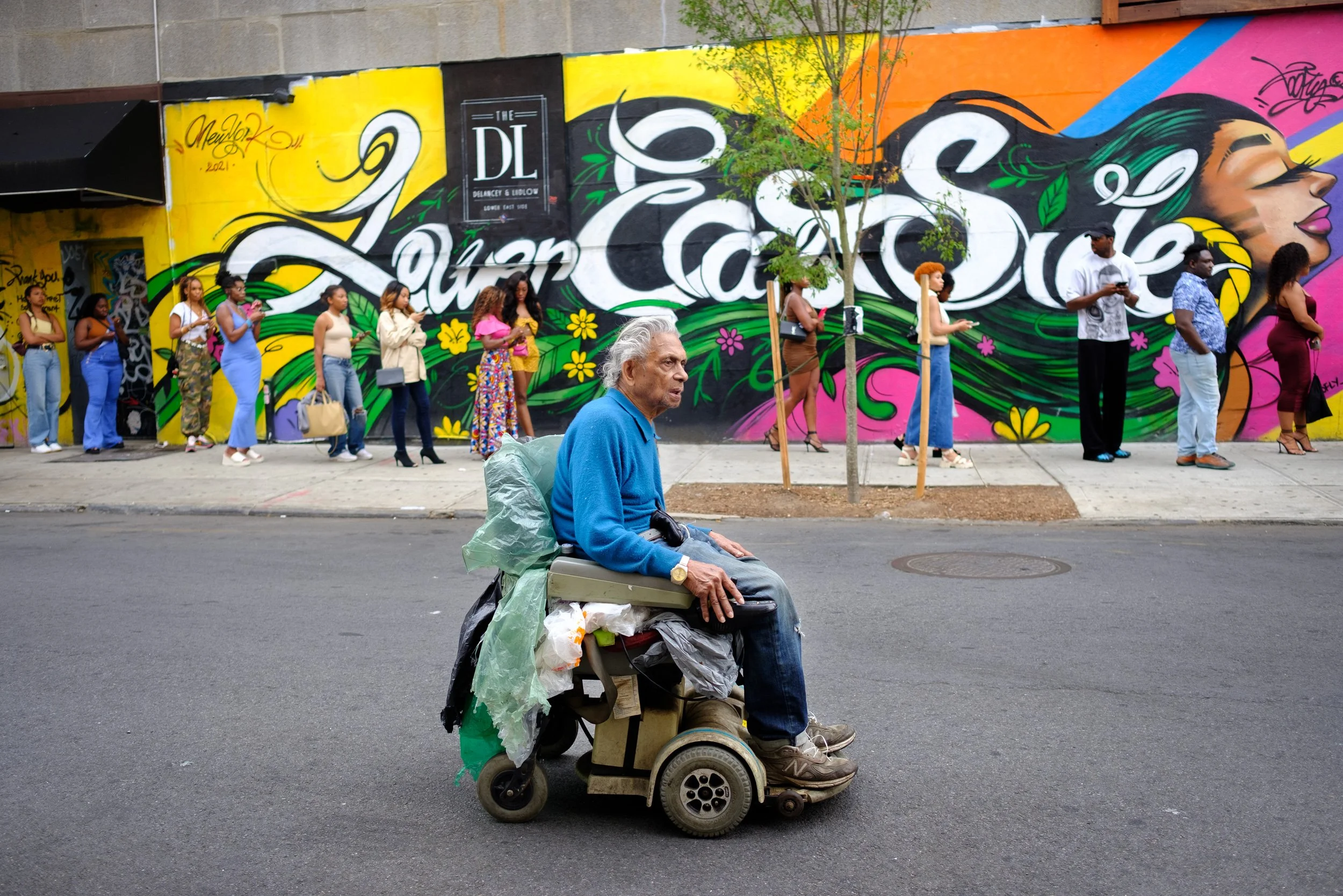 An elderly man with gray hair sitting in a motorized wheelchair on a city street. He has a blue sweater and is surrounded by bags and trash. In the background, a group of people including women and men stands in line in front of a colorful mural on a