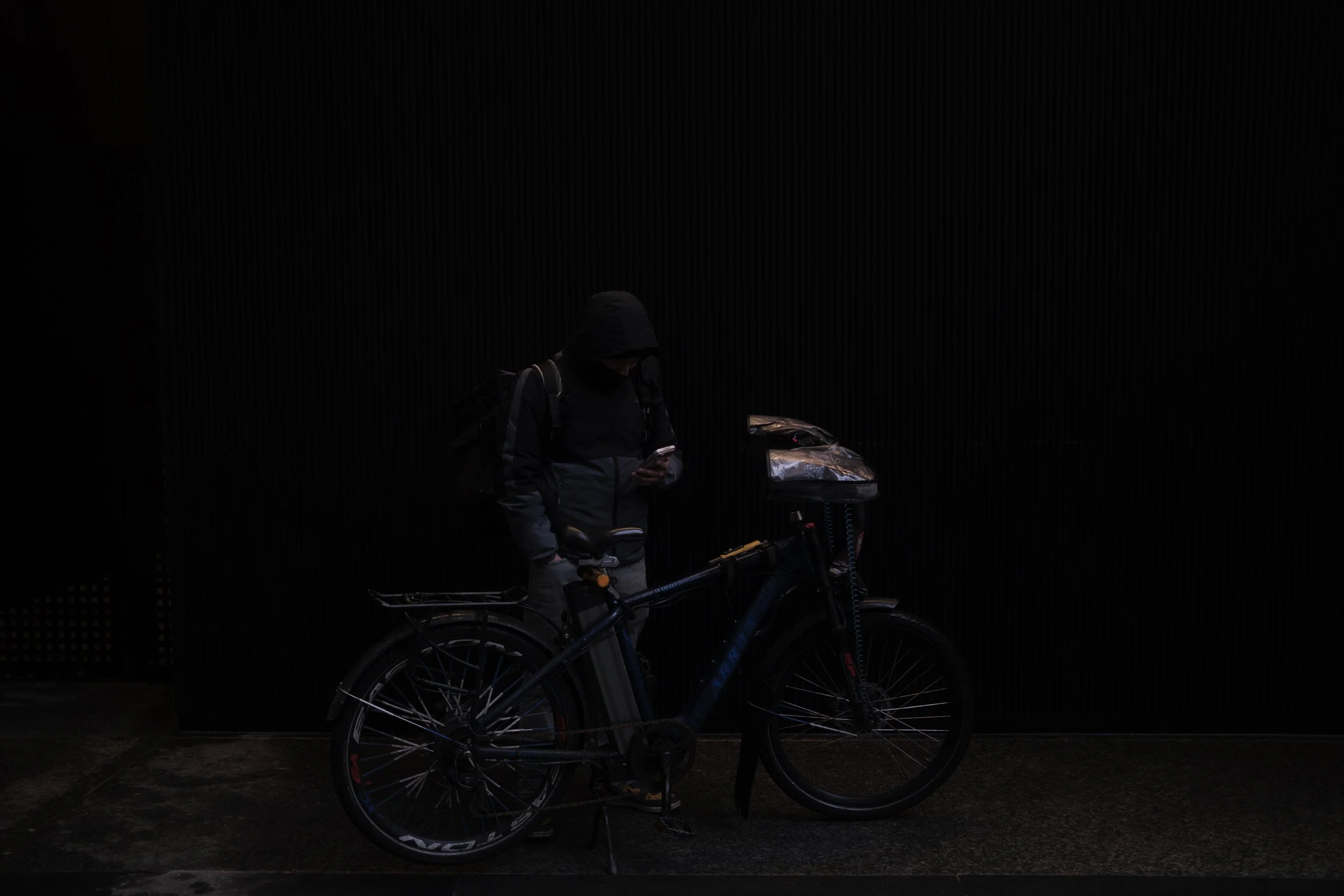 A person in dark clothing, hooded, standing next to a bicycle and looking at their phone, against a dark, textured wall.