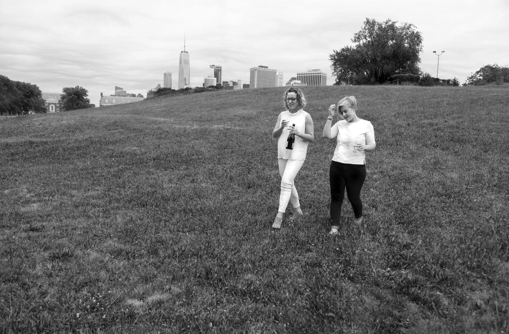 Two women walking on grass in a park with city skyline in the background, one holding a bottle and the other touching her hair.