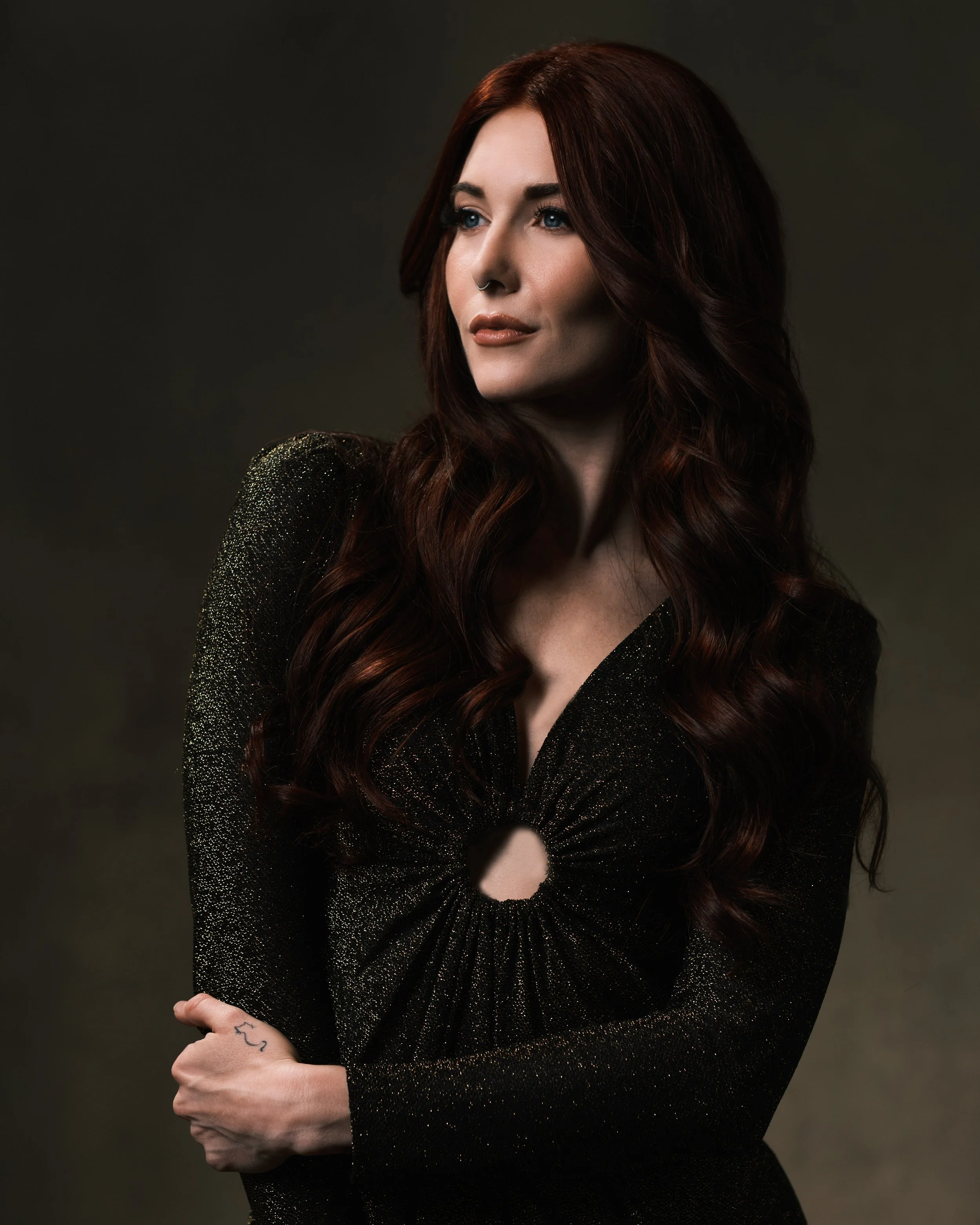 A woman with long auburn wavy hair, wearing a black, shimmering, long-sleeve dress with a cutout at the waist, is posing against a dark, blurred background. She has light makeup, blue eyes, and a septum piercing, with her left arm crossed over her body.