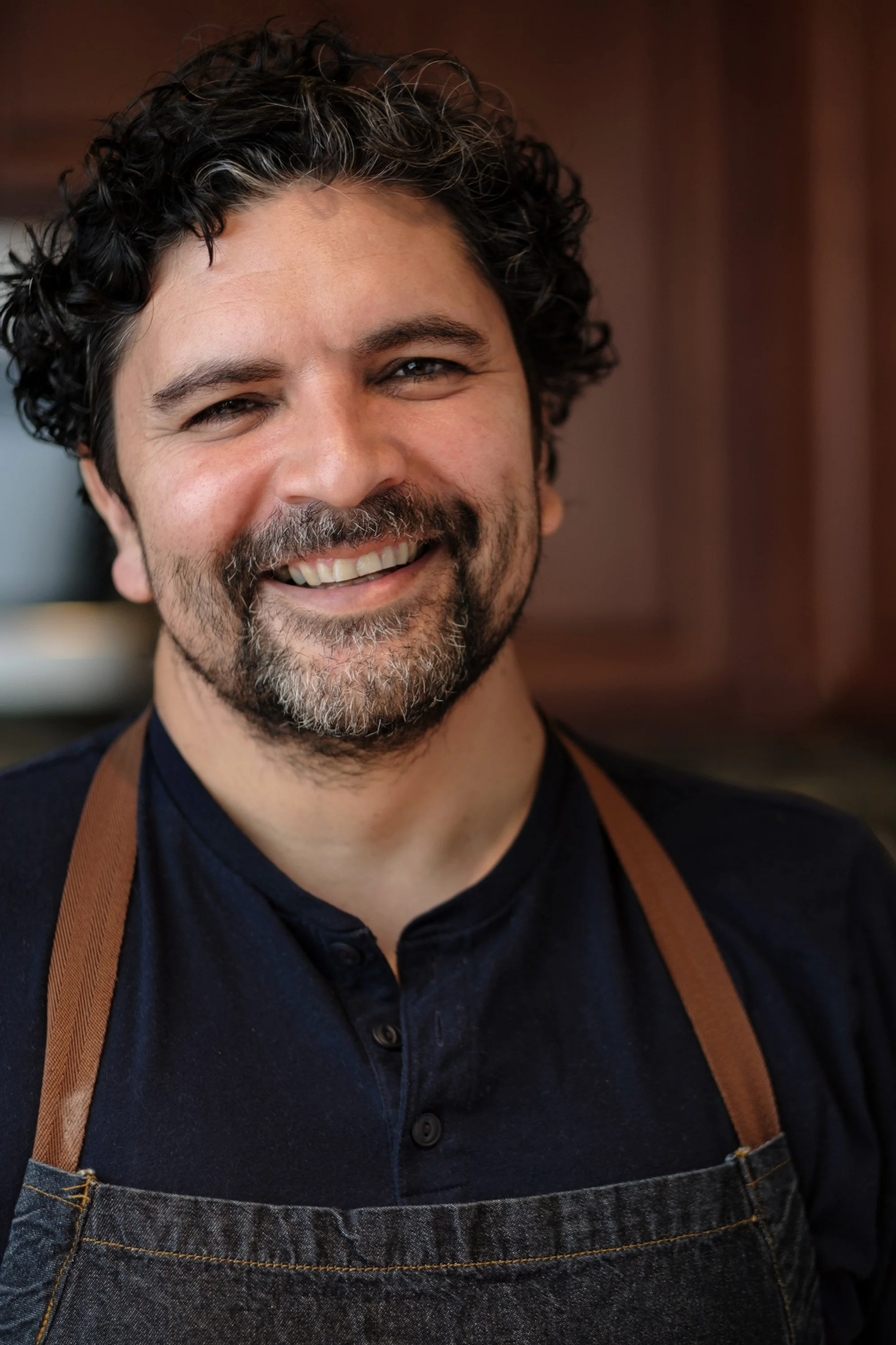 A smiling man with curly dark hair and a beard, wearing a black shirt and denim apron, with a brown strap from an apron or bag over his shoulder, in front of a blurred wooden background.