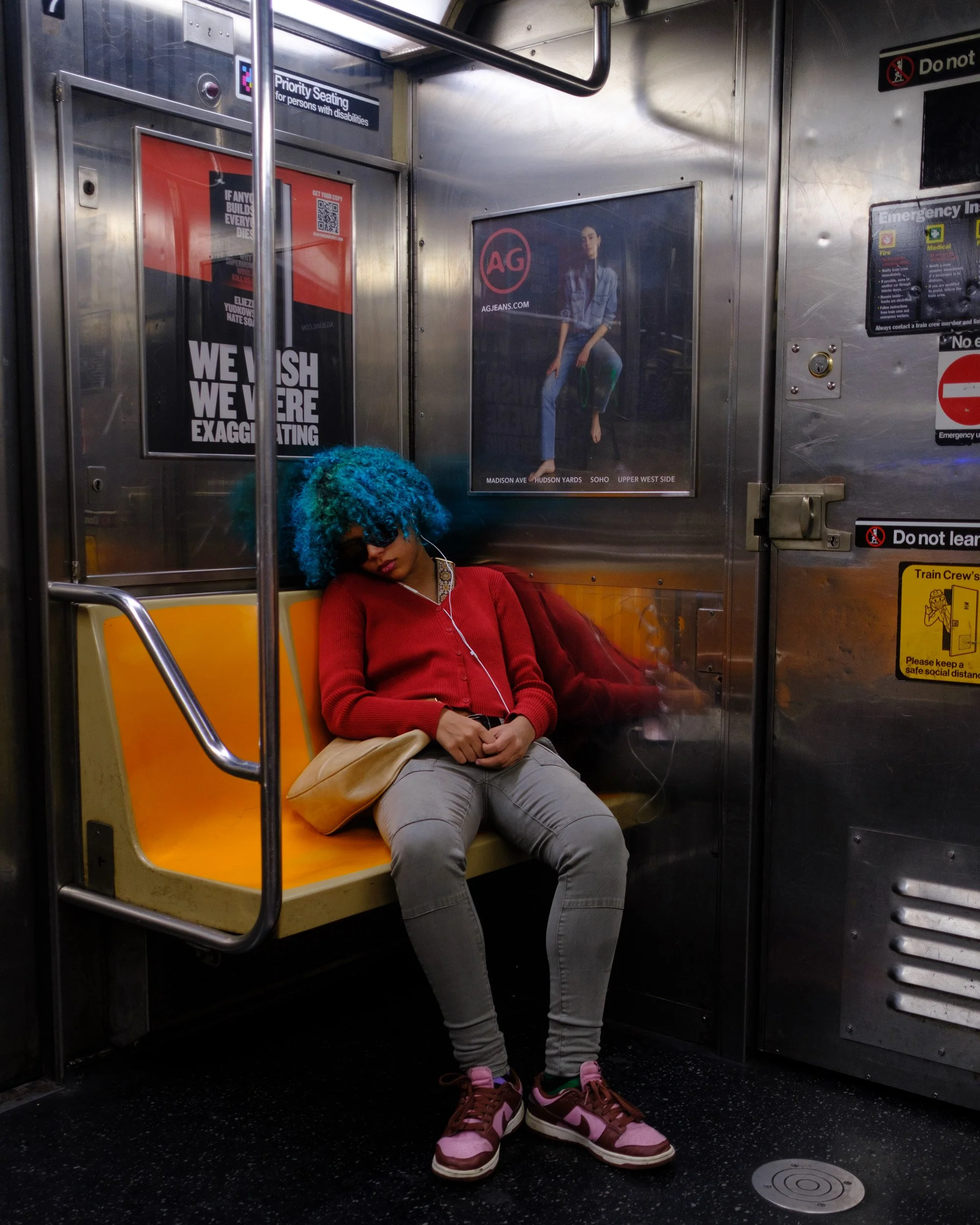 A woman with curly blue hair, beige pants, a red sweater, and pink and brown sneakers is sitting on an orange metro train seat with her eyes closed, listening to music with earphones.