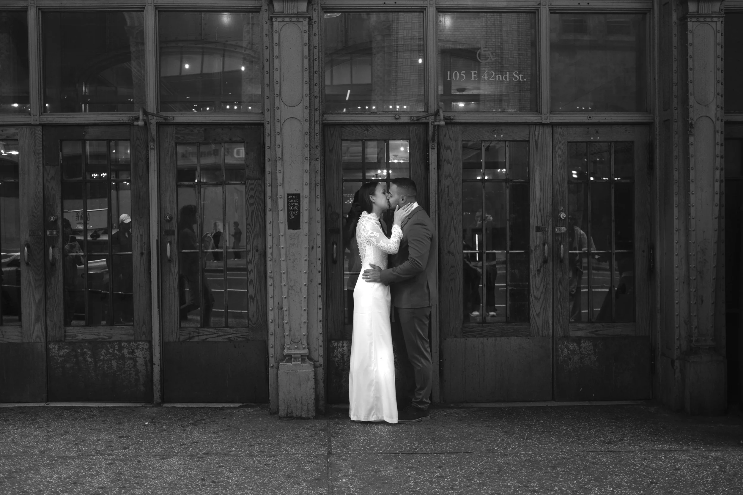 A couple in wedding attire kissing in front of a glass and wood door at night.