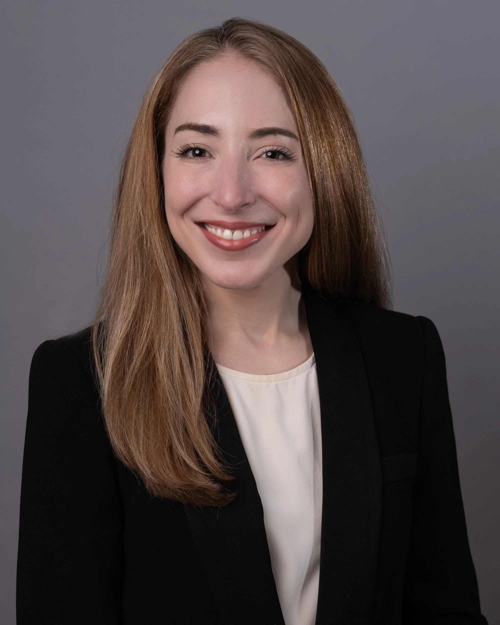 A woman with long, straight, red hair smiling and wearing a black blazer with a white top, posed against a plain gray background.
