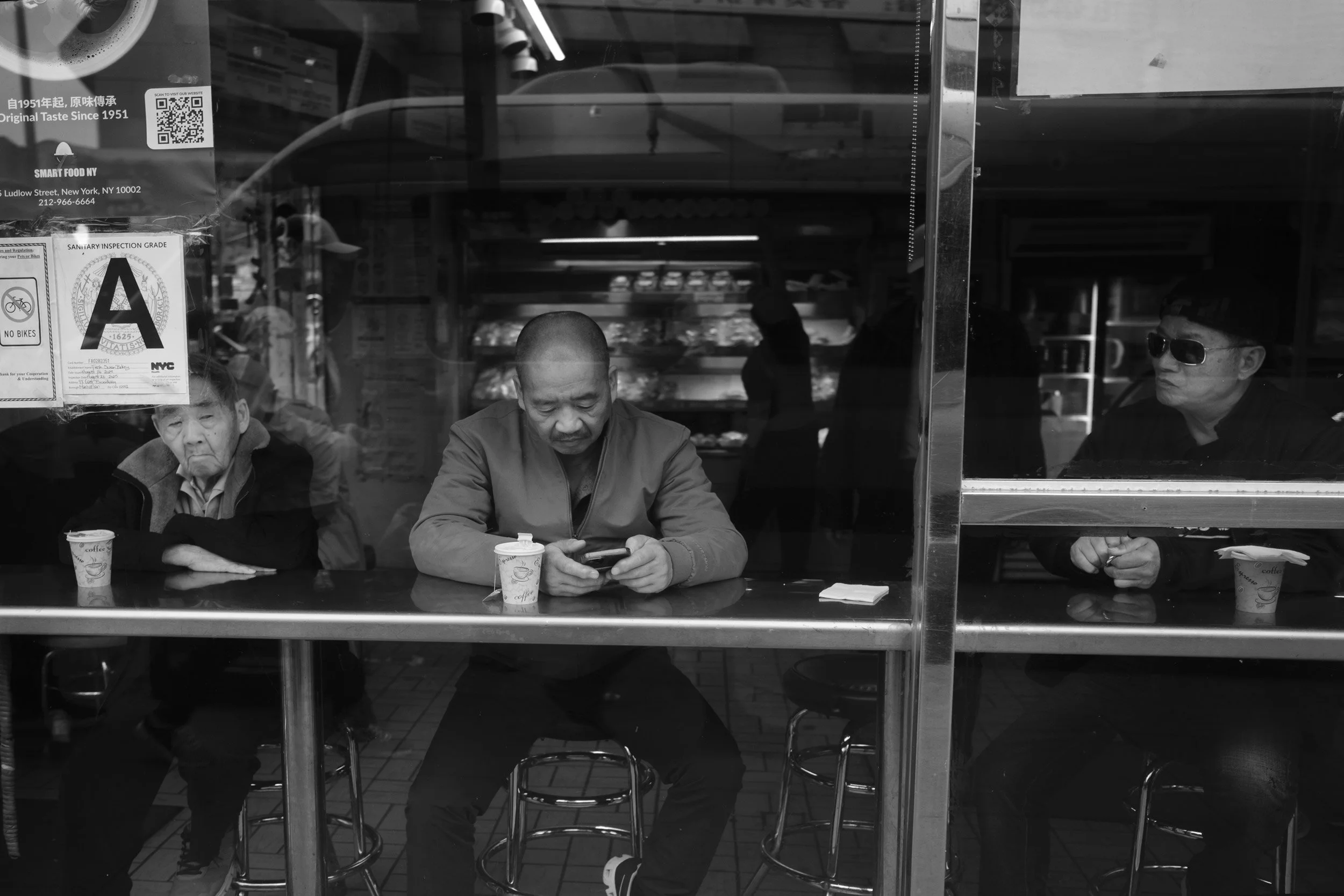 Three men sitting at a counter inside a fast-food restaurant, with two of them engaged with their phones and the third looking down at a cup. The scene is viewed through a reflection on the window, creating a layered effect.