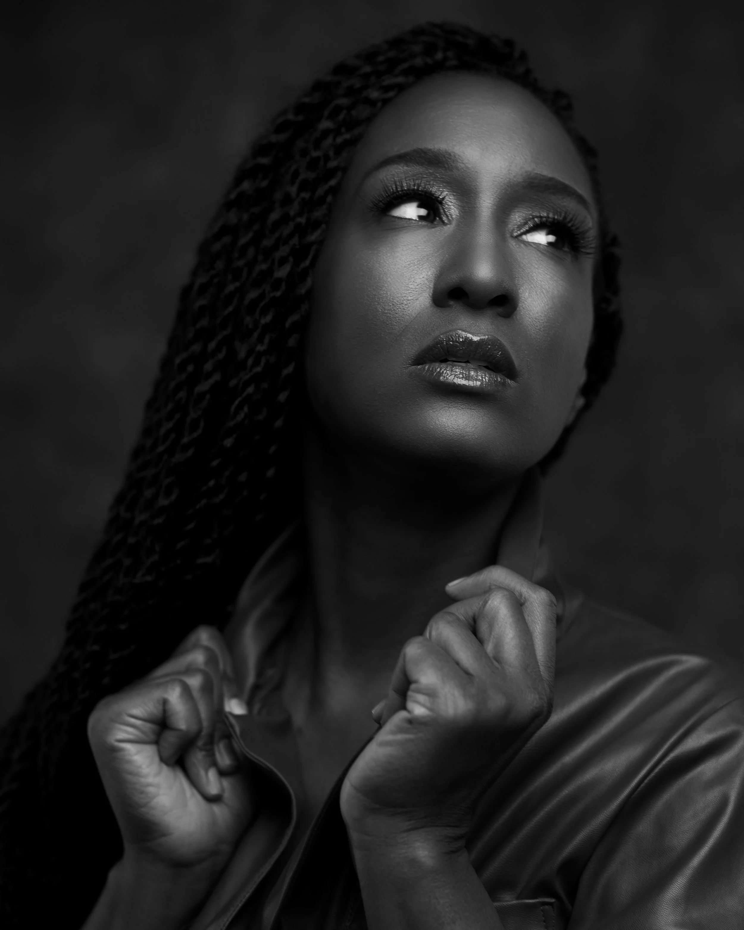 Black and white photo of a woman with braided hair, intense eyes, and glossy lips, looking upward with hands clenched, near her chest.