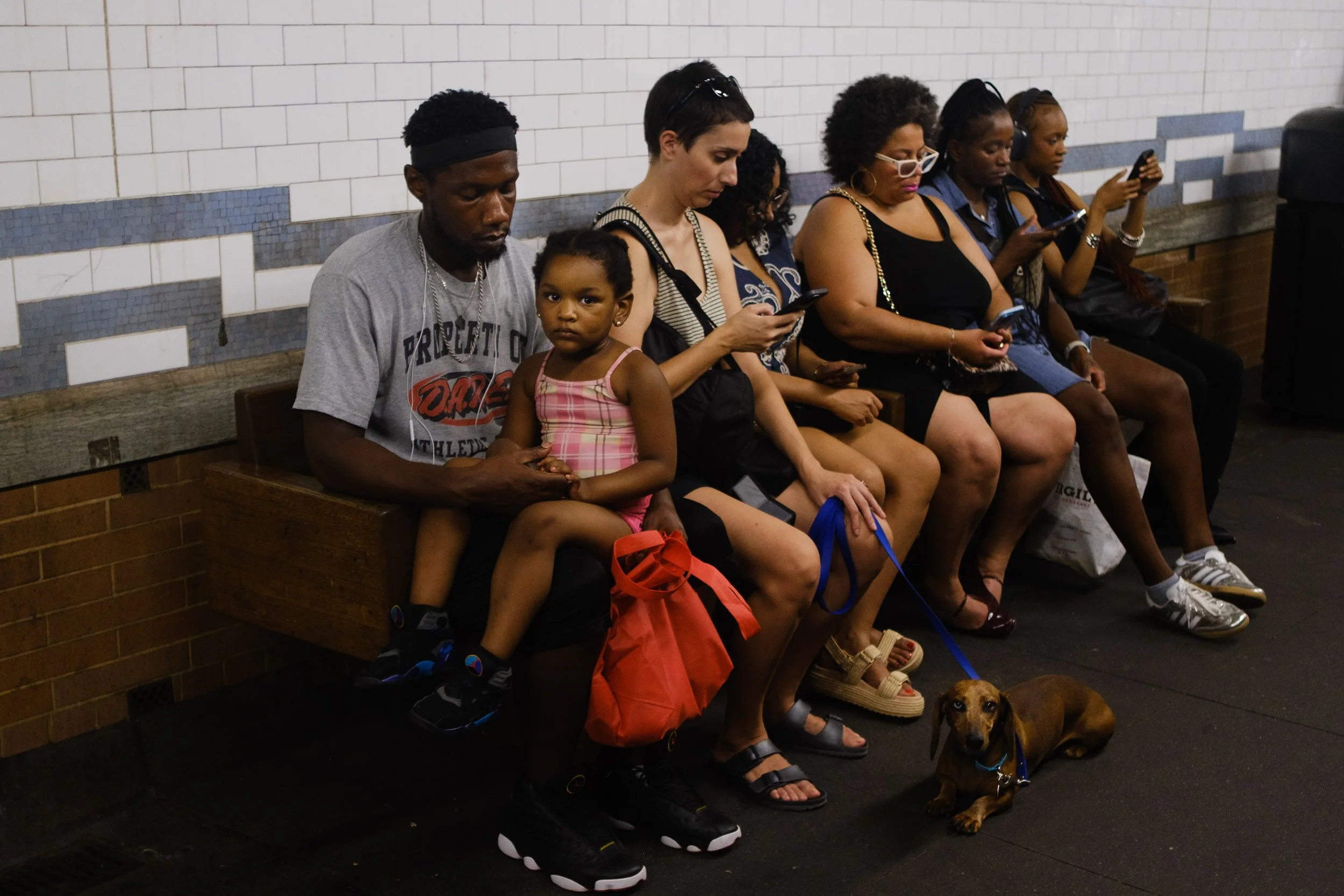 People sitting on a bench waiting, with a dog on a leash at their feet, in a subway or train station. They are focused on their phones and wearing casual clothing.