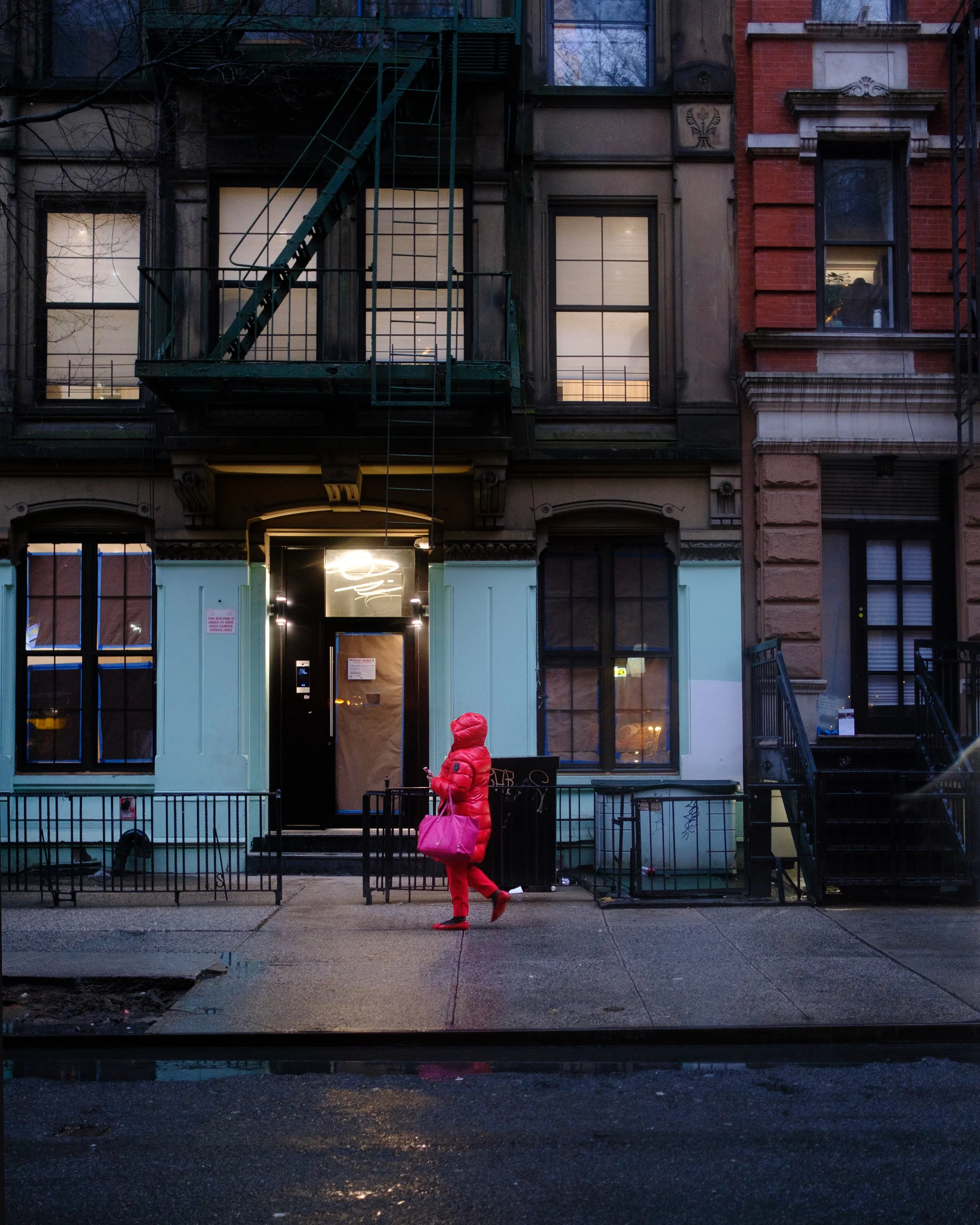 A person in a bright red coat and pants walking on a city sidewalk at dusk or dawn, carrying a pink bag. There are old brick and stone buildings with lit windows and fire escape stairs in the background.