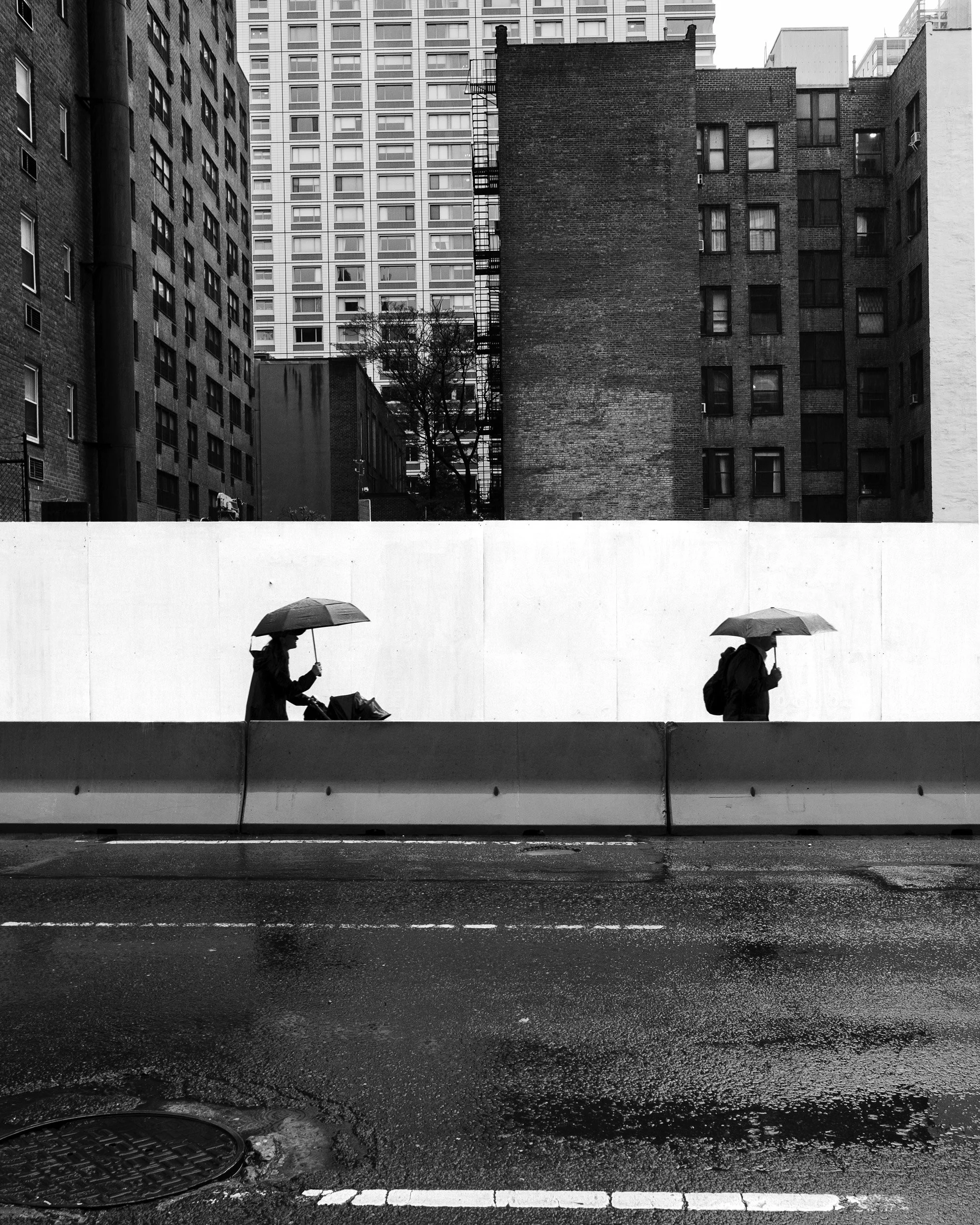 Black and white photograph of a city scene with a white concrete barrier in the foreground, two people walking with umbrellas, several tall buildings, and leafless trees in the background.