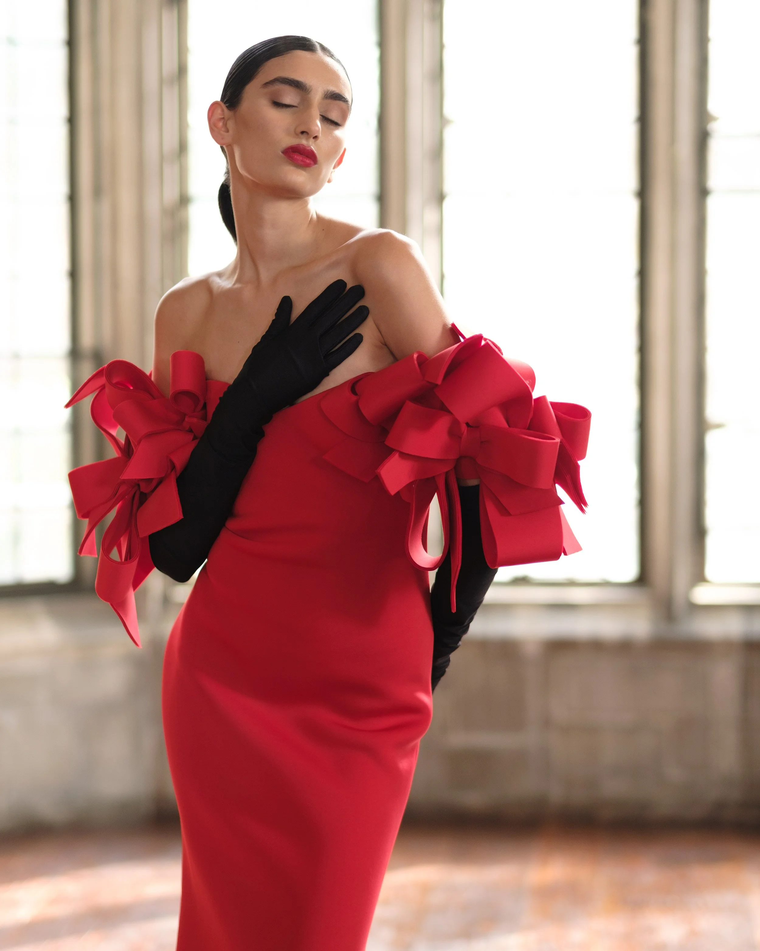 A woman in a red dress with large bow details, wearing black gloves, posing with her eyes closed and hand on her shoulder in front of large windows.