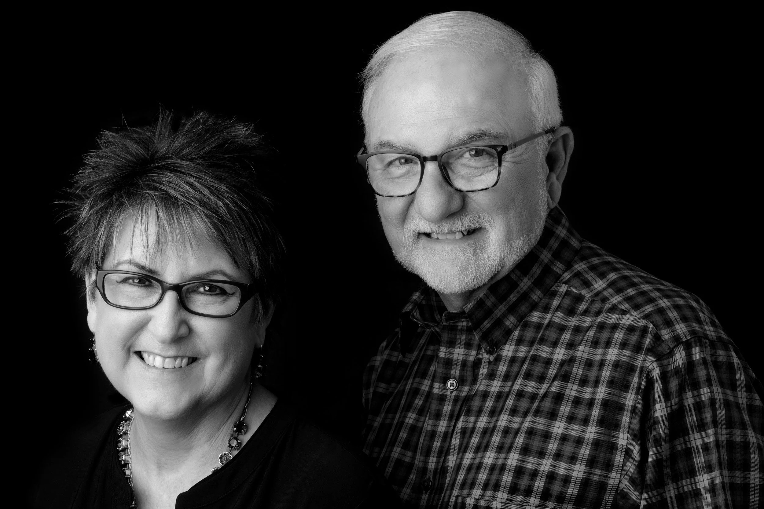 Black and white portrait of an older woman wearing glasses, smiling, with short curly hair and a necklace, next to an older man with glasses, smiling, with short light-colored hair, a beard, and wearing a plaid shirt.