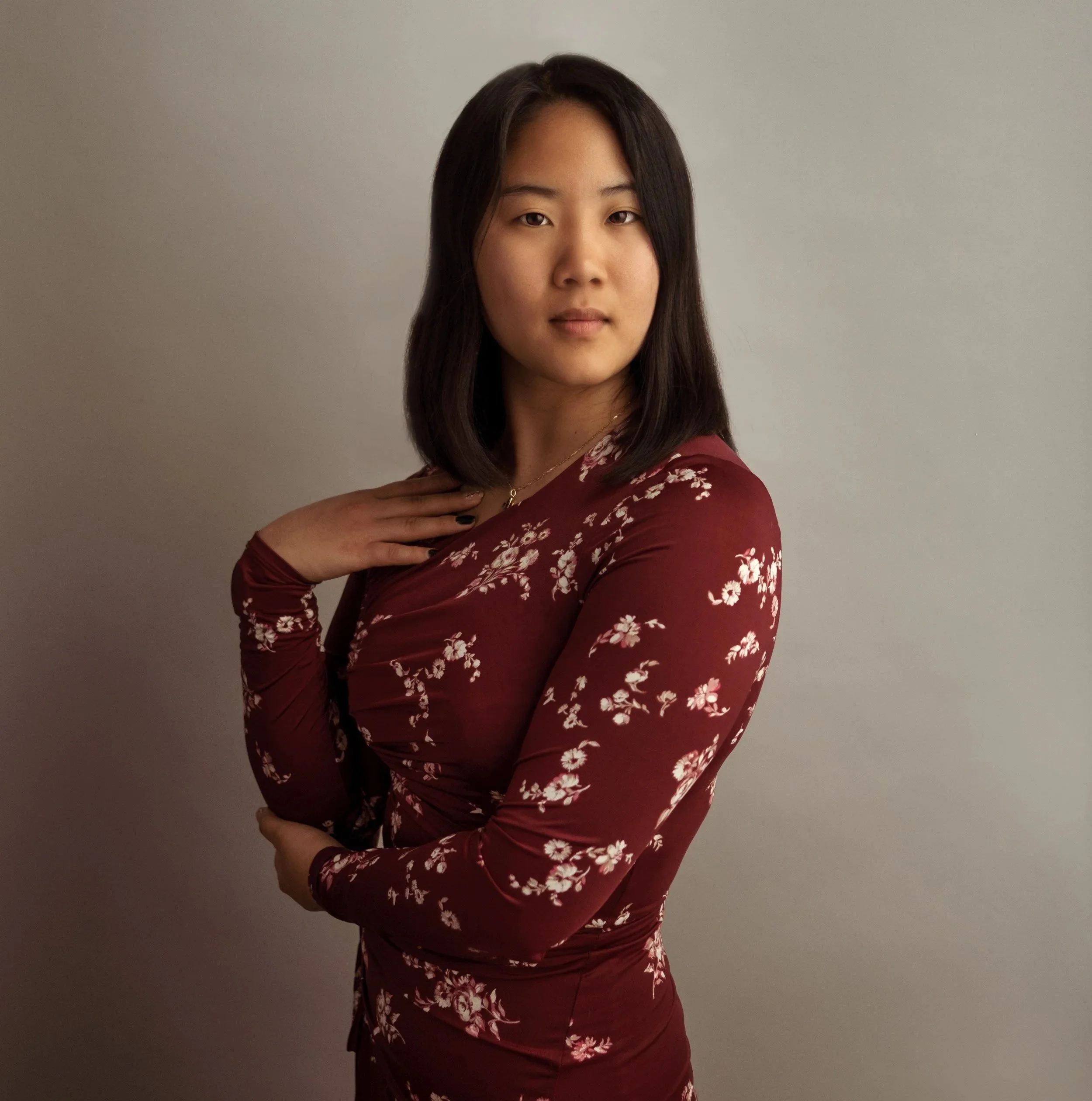 A young woman with long dark hair wearing a red floral long sleeve top, posing with her right hand on her shoulder, against a plain gray background.