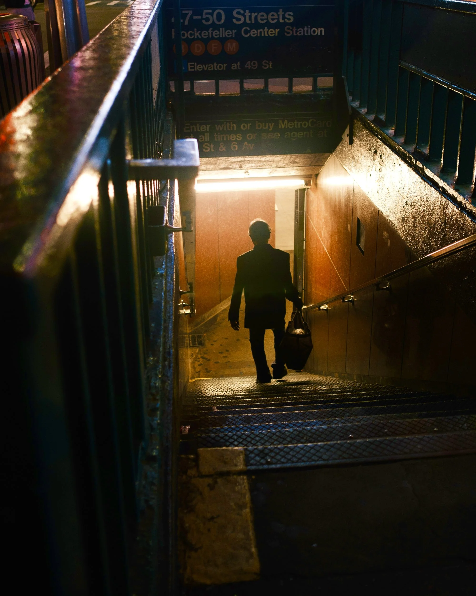 Person ascending an underground subway entrance staircase at night, holding a bag. The subway entrance sign indicates it is for the 1, 7, 50 Streets stations near Rockefeller Center, with instructions to enter with a MetroCard and directions to the e