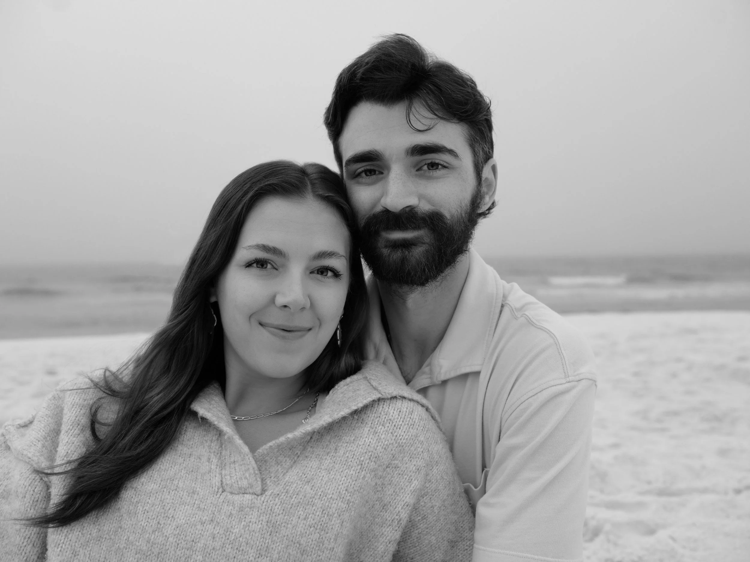 A black and white photo of a couple at the beach, close-up shot of their faces showing a woman with long hair and a man with a beard, both smiling softly.
