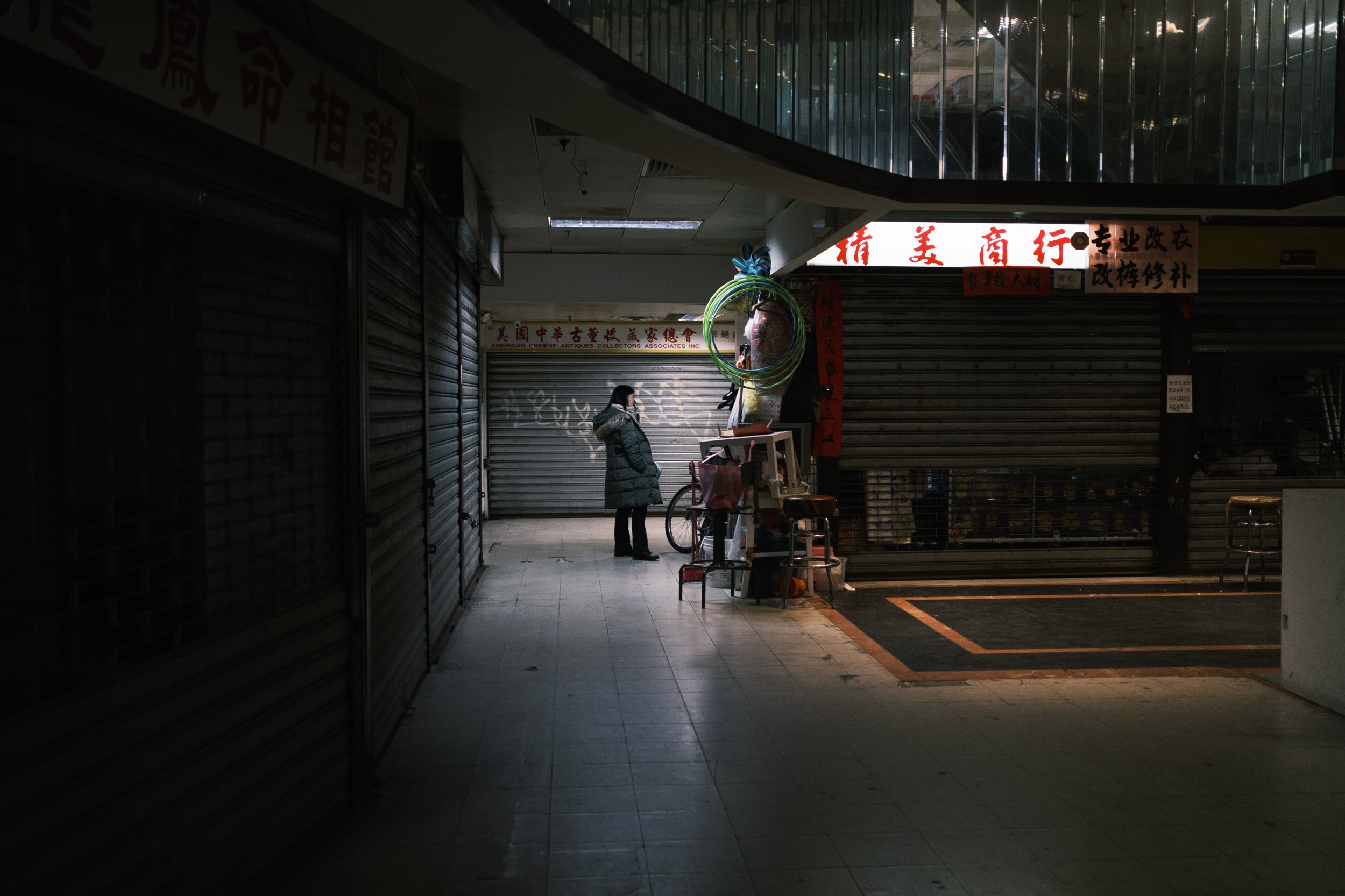 A woman in a coat standing outside a closed shop with metal shutters in an urban area at night. The shop has Chinese characters on signs and a few items like balloons and a bicycle nearby.