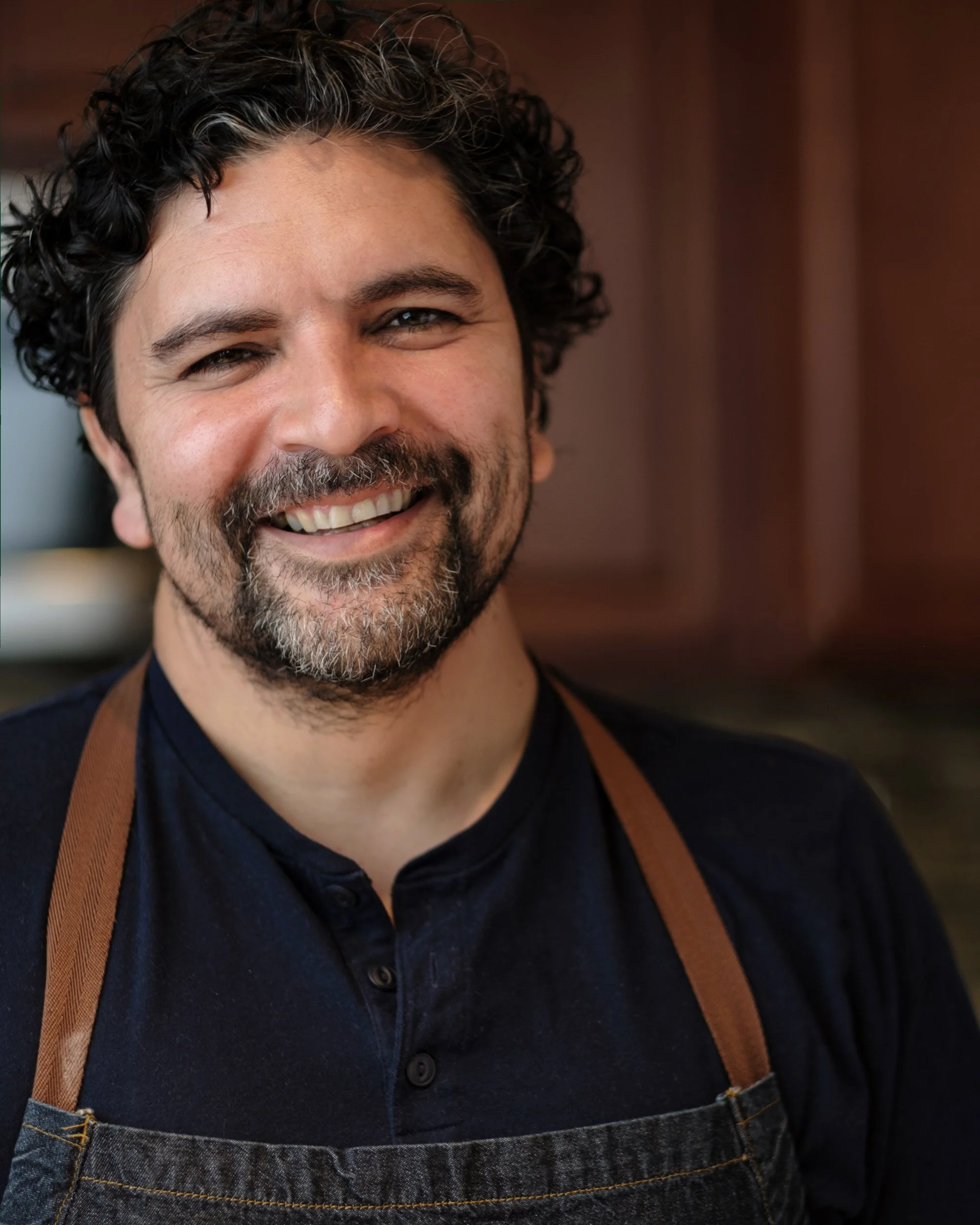 Close-up of a smiling man with curly dark hair, a beard, wearing a dark shirt, an apron, and a brown strap over his shoulder.
