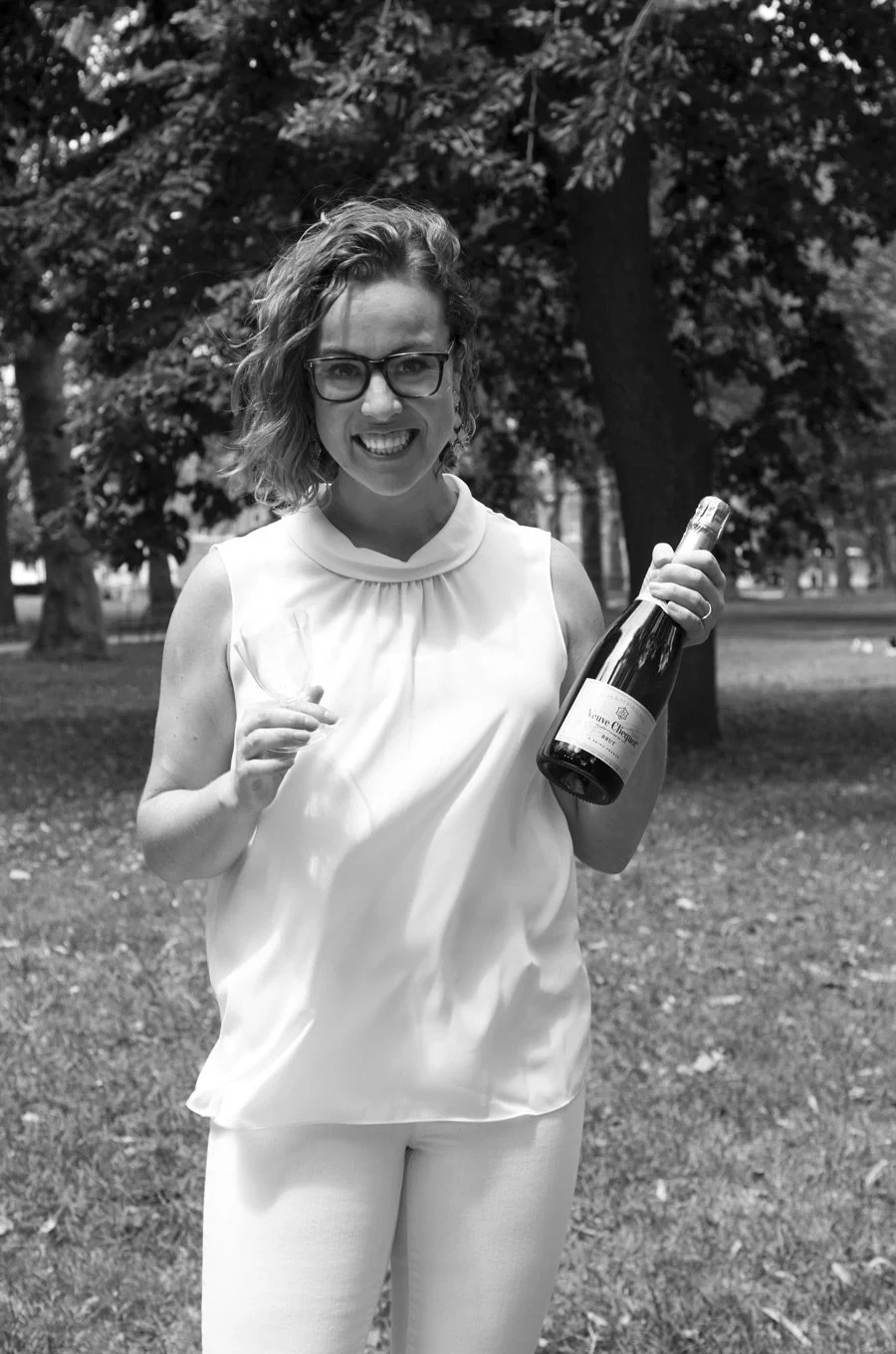 A woman with glasses and curly hair smiling while holding a wine bottle and a glass, standing outdoors in a park with trees.