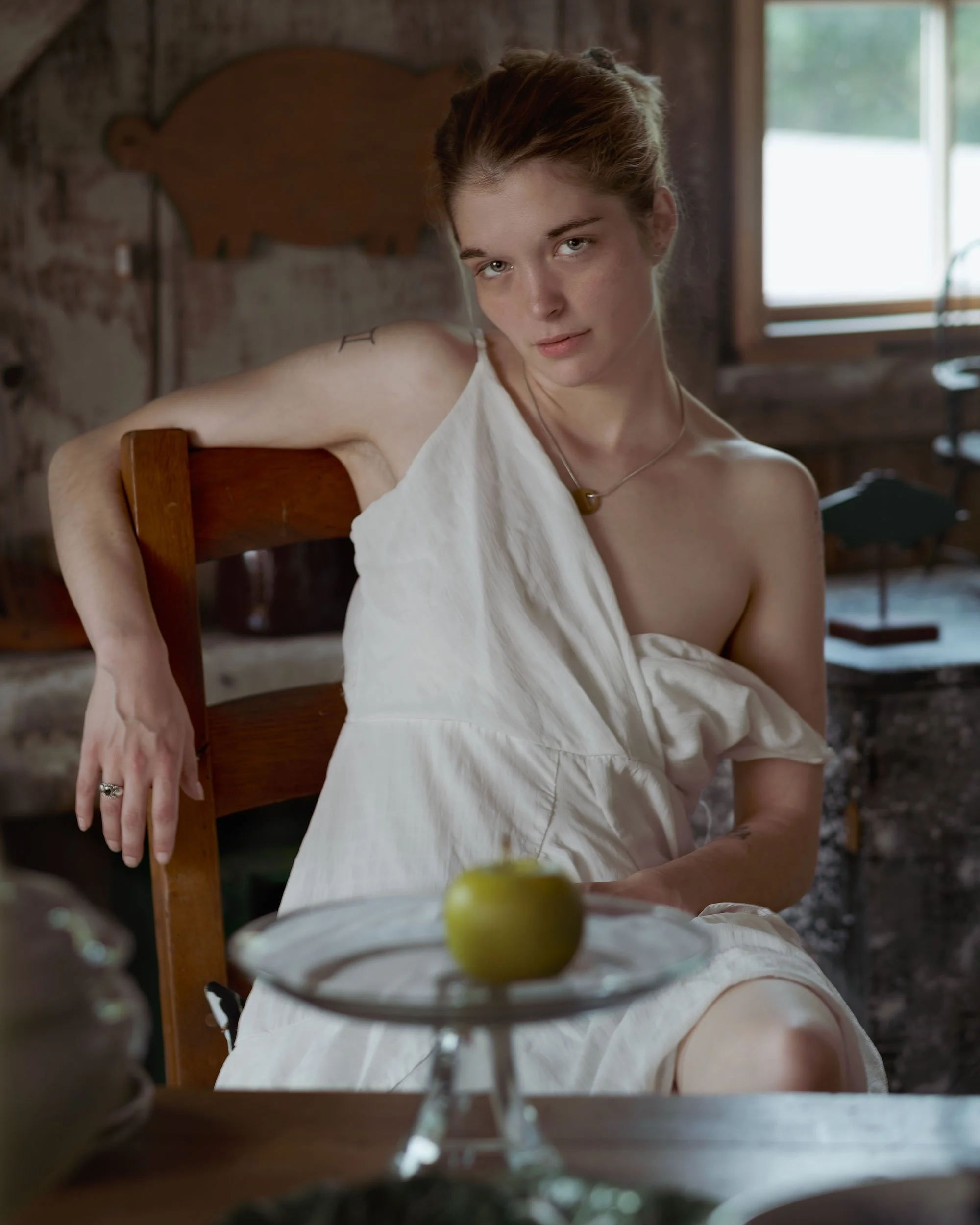 A woman with reddish-brown hair and light skin sitting in a wooden chair in a rustic room, wearing a white sleeveless dress and a necklace, looking at the camera with a neutral expression. A glass with a lime garnish is on the table in front of her, 