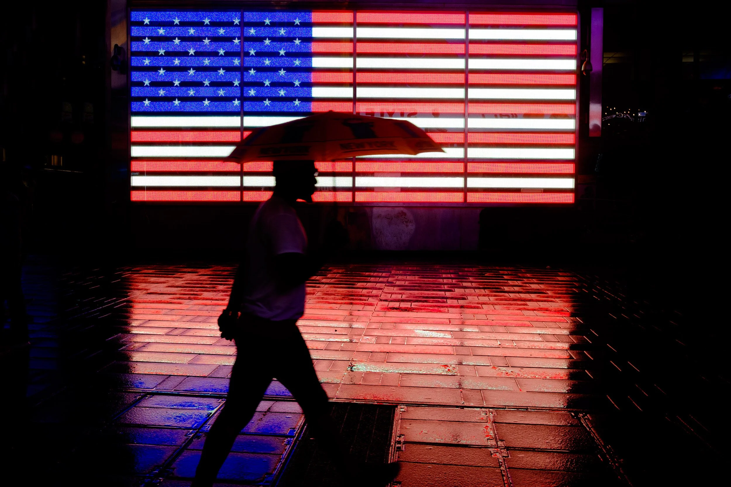 Silhouette of a person walking with an umbrella in front of an illuminated American flag display at night.
