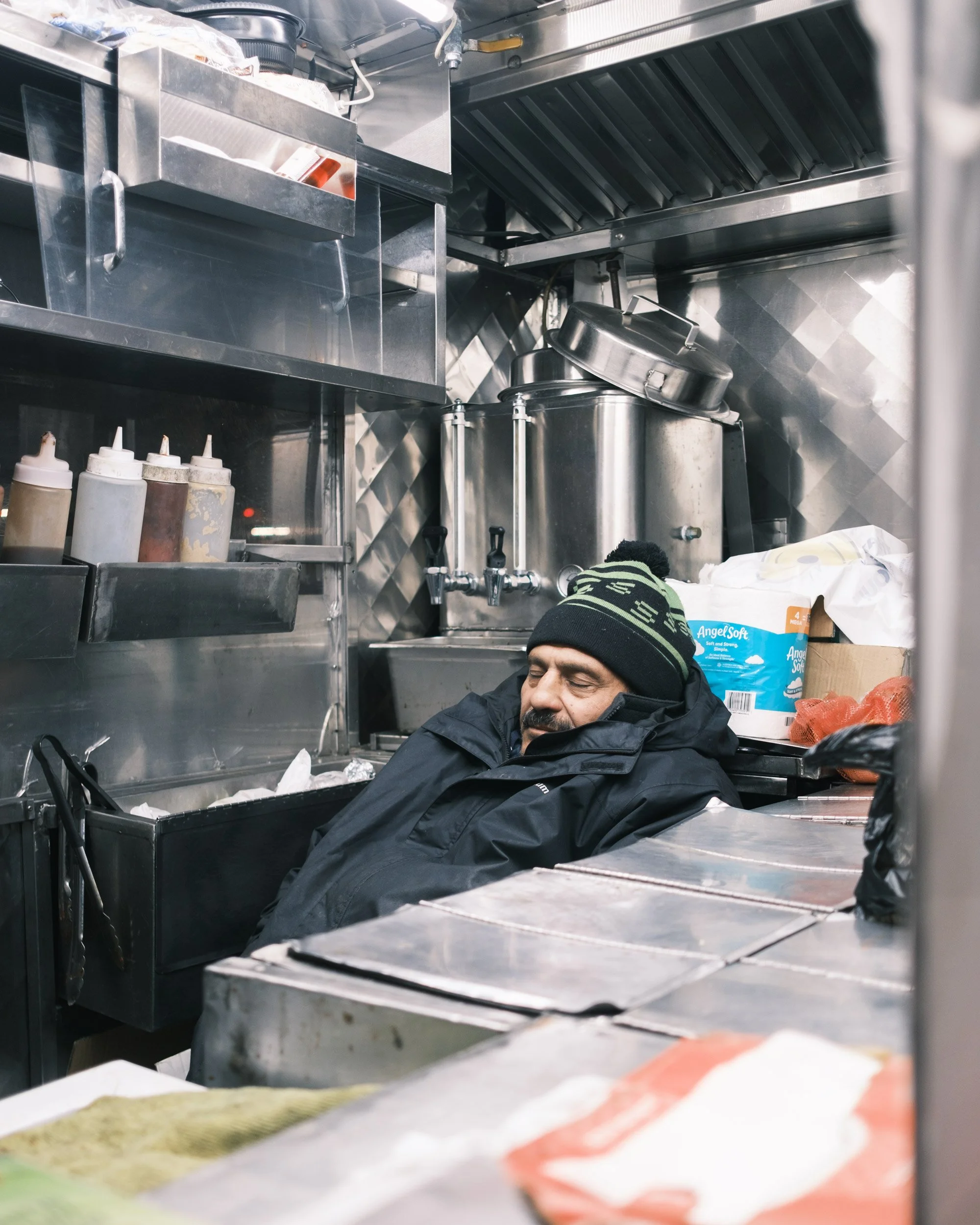 A man sleeping in a commercial kitchen with stainless steel surfaces, shelves, and kitchen equipment around him.