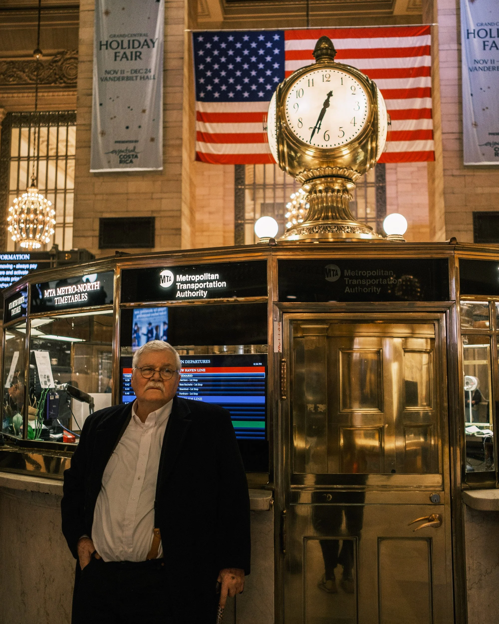 Interior of a train station with a large clock on top of a metal structure, displaying the time as 6:02. An American flag and a white banner advertising a holiday fair are visible in the background. A man with glasses in a suit stands in front of the