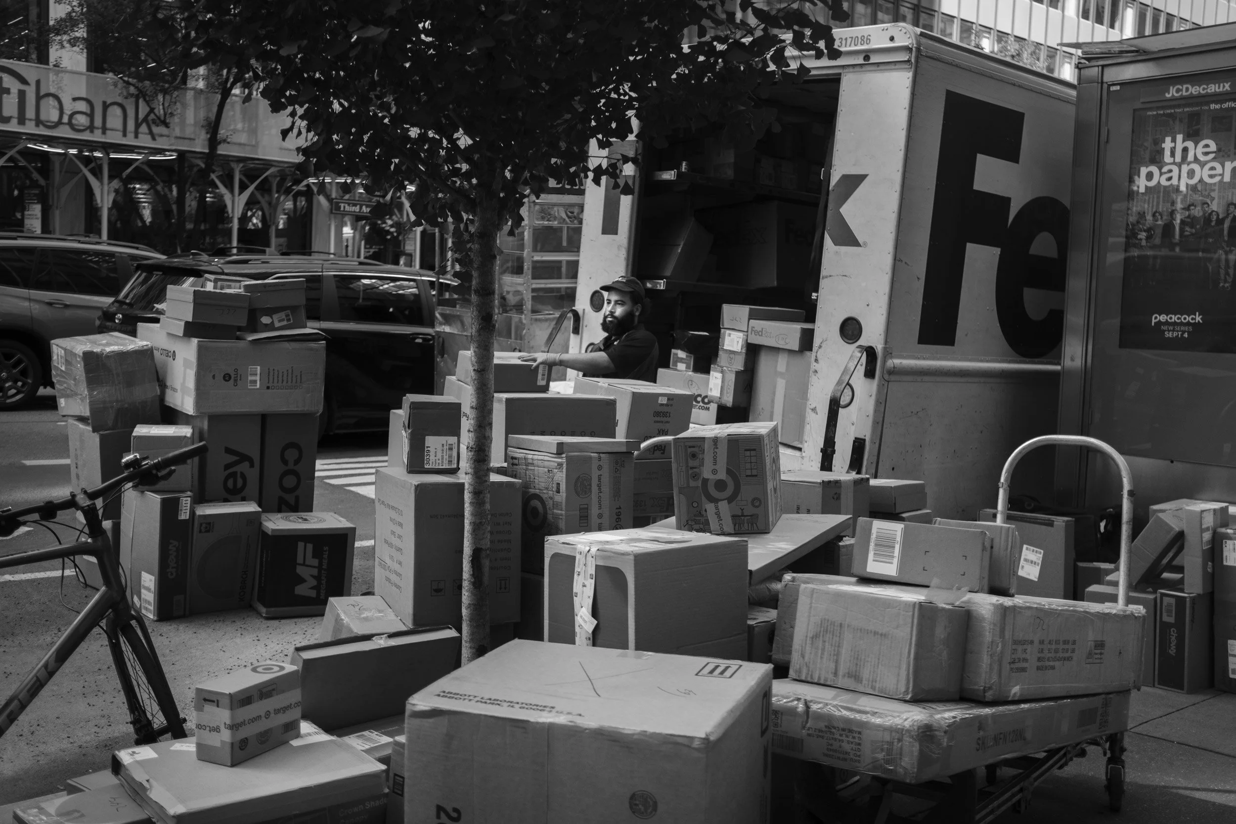 A man with a beard and cap loading boxes into a FedEx truck on a city street, with cars parked nearby and a tree in the foreground.