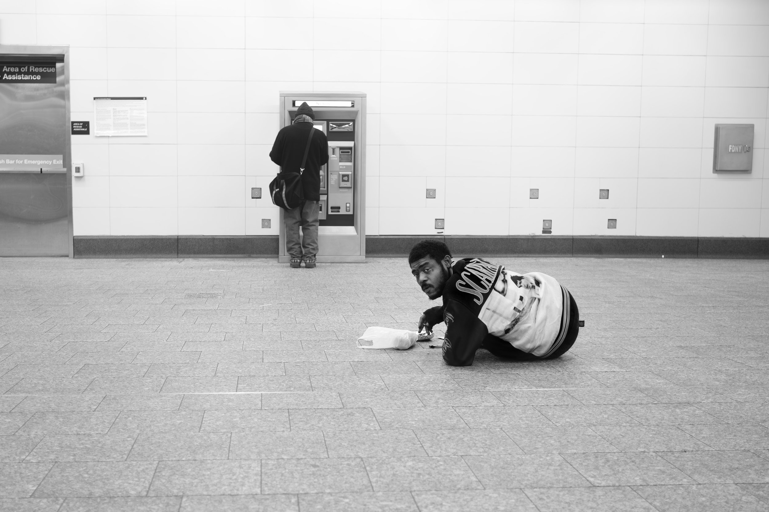 A person with a hood and backpack standing at a vending machine, and another person lying on the floor with a plastic bag.
