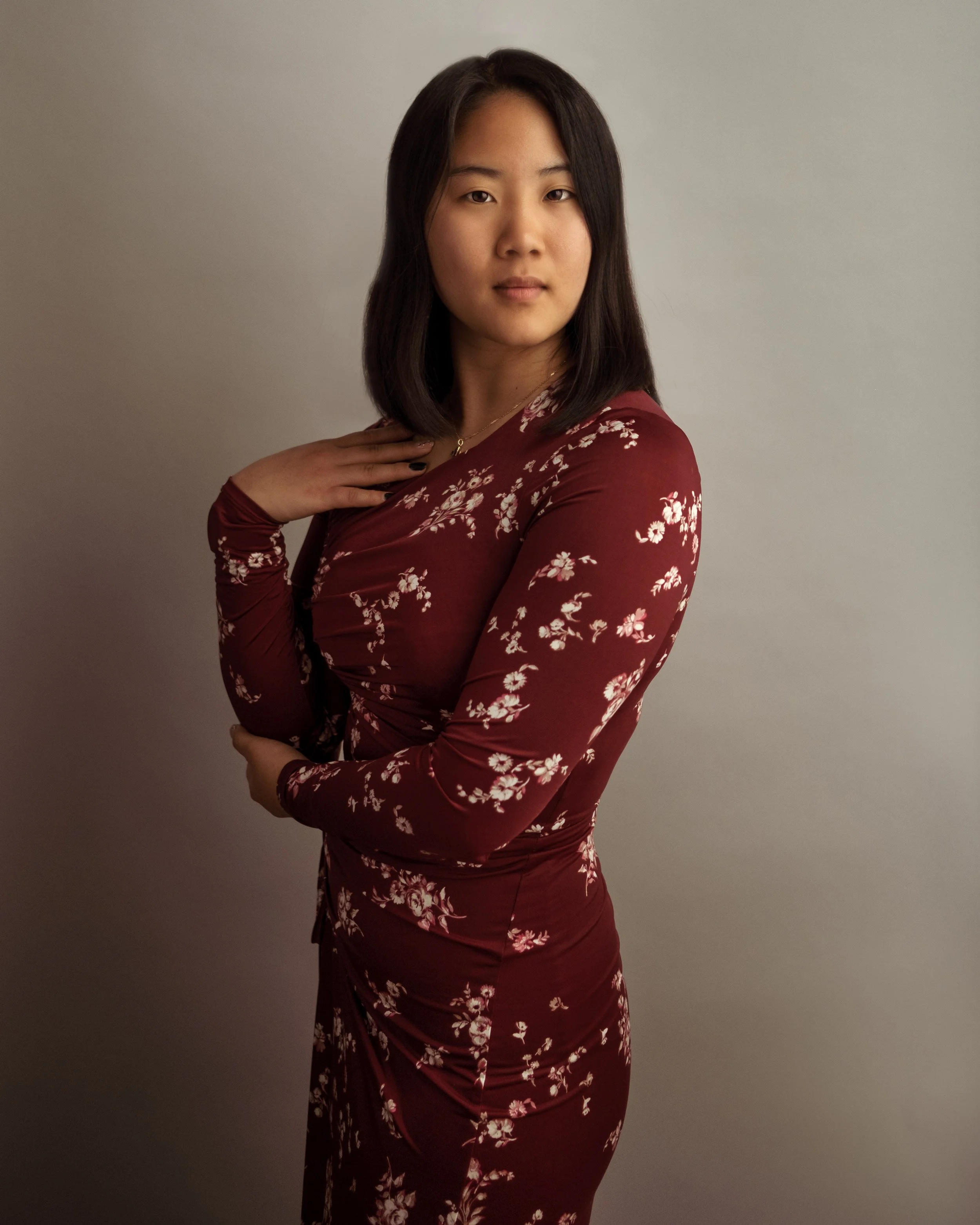 A young woman with medium skin tone and dark brown hair, wearing a burgundy dress with white floral pattern, standing against a plain gray background.