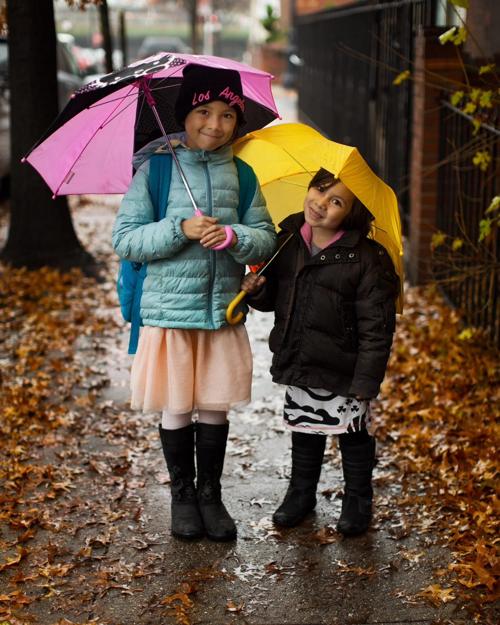 Two young girls walking outside on a wet, leaf-covered sidewalk, holding colorful umbrellas and smiling, dressed warmly for the rainy weather.