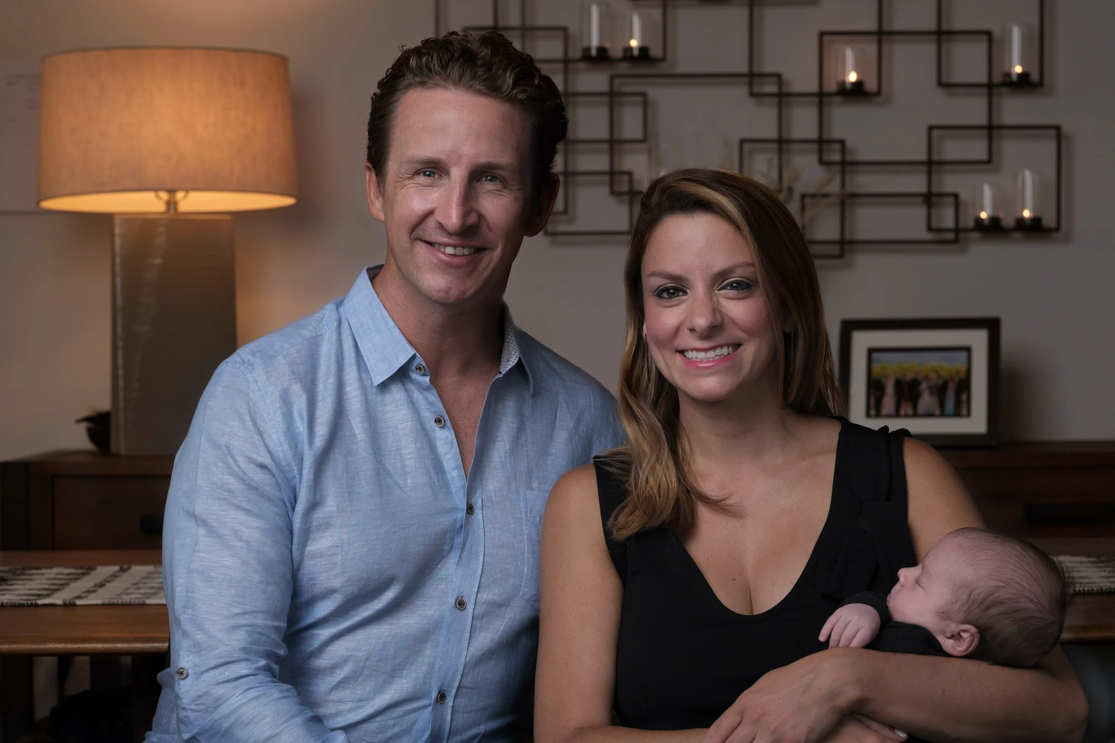 A smiling man in a light blue shirt standing next to a smiling woman with long wavy hair holding a baby, inside a home with decorative wall shelving and lit candles in the background.