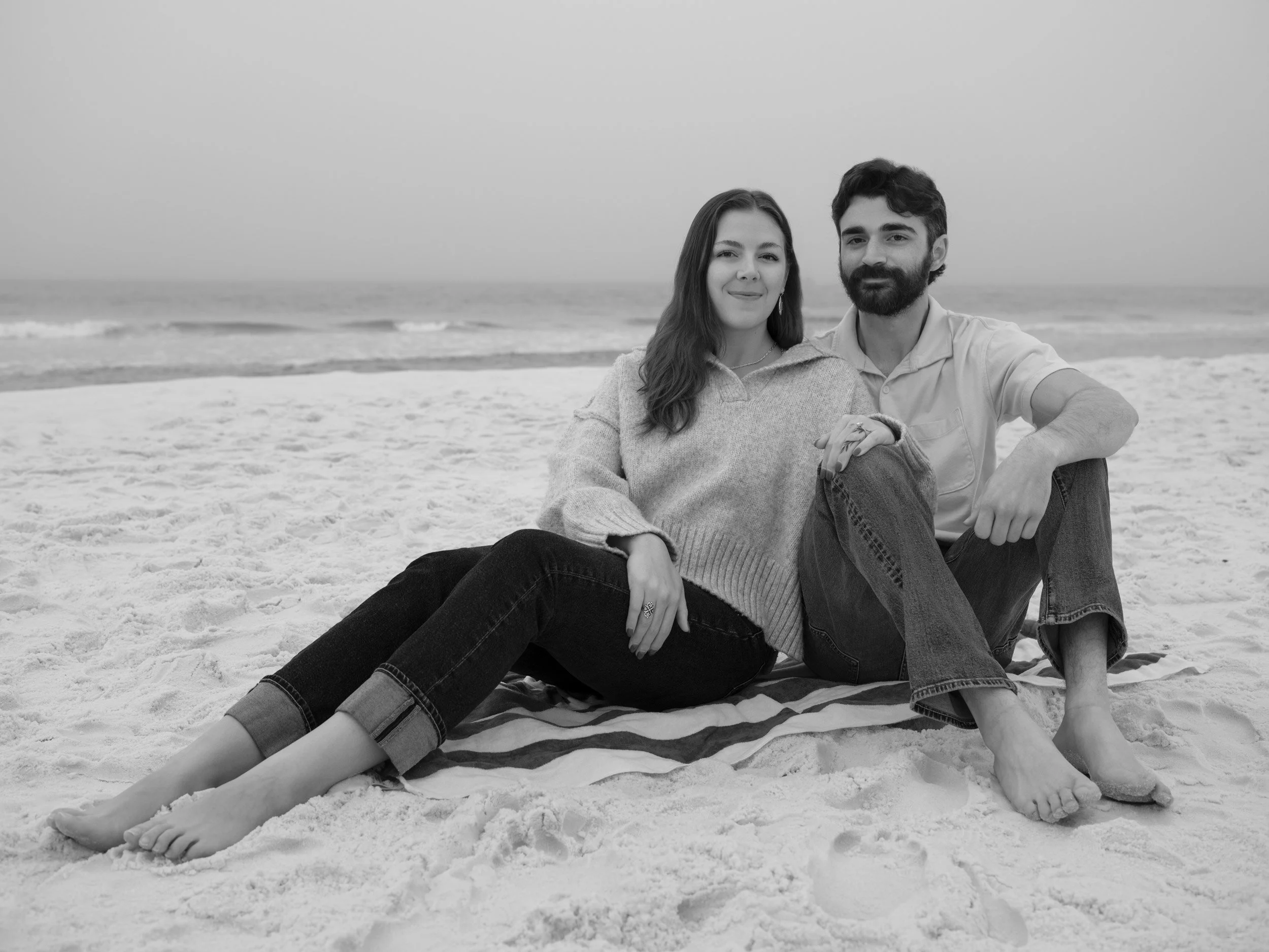 A black and white photo of a young couple sitting on the sand at the beach, smiling at the camera. The woman has long hair and is wearing a sweater, while the man has a beard and is dressed in a shirt. They are sitting close together with the ocean w