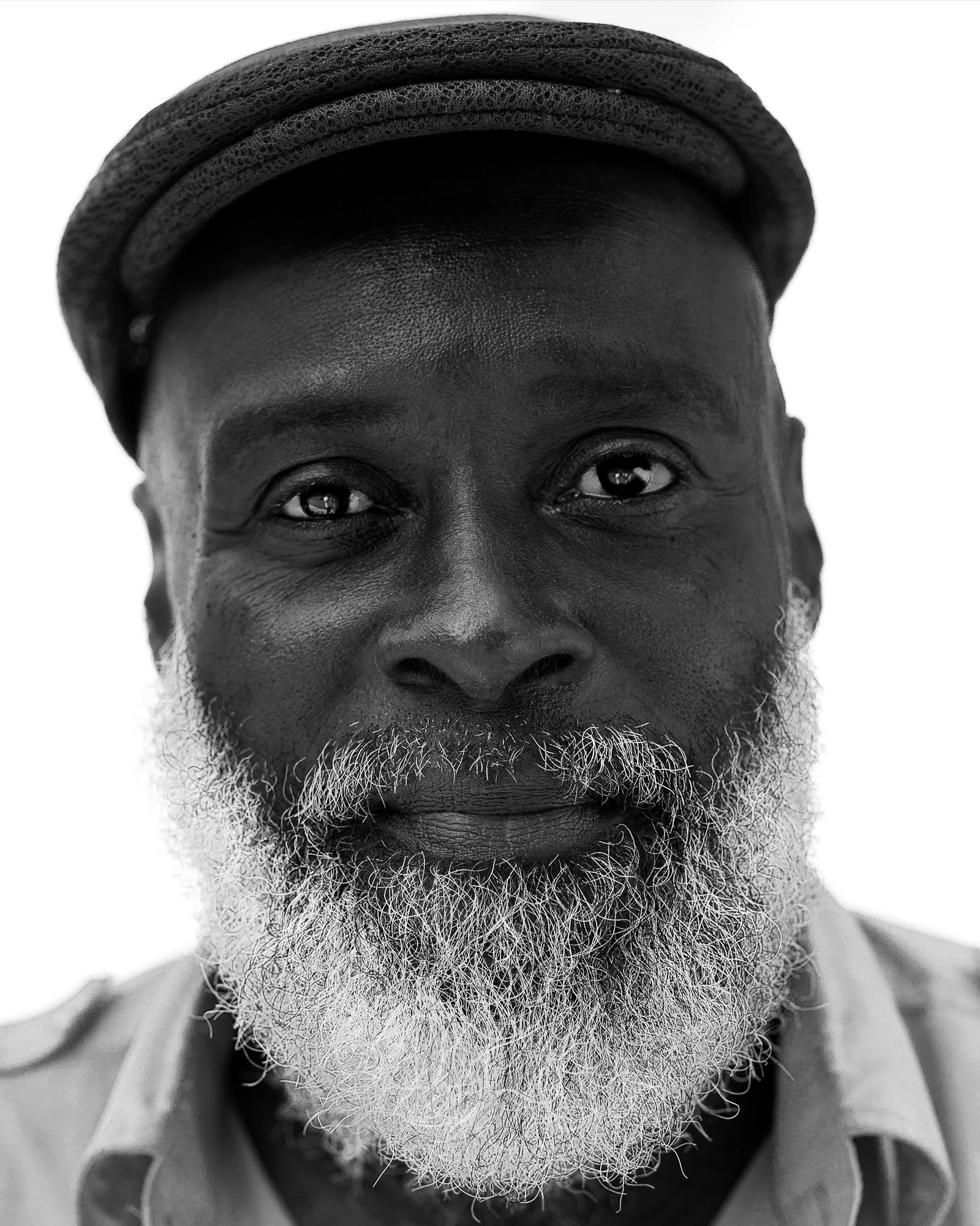 Close-up black-and-white photo of a man with a beard wearing a cap, with his face filling the frame and a plain white background.