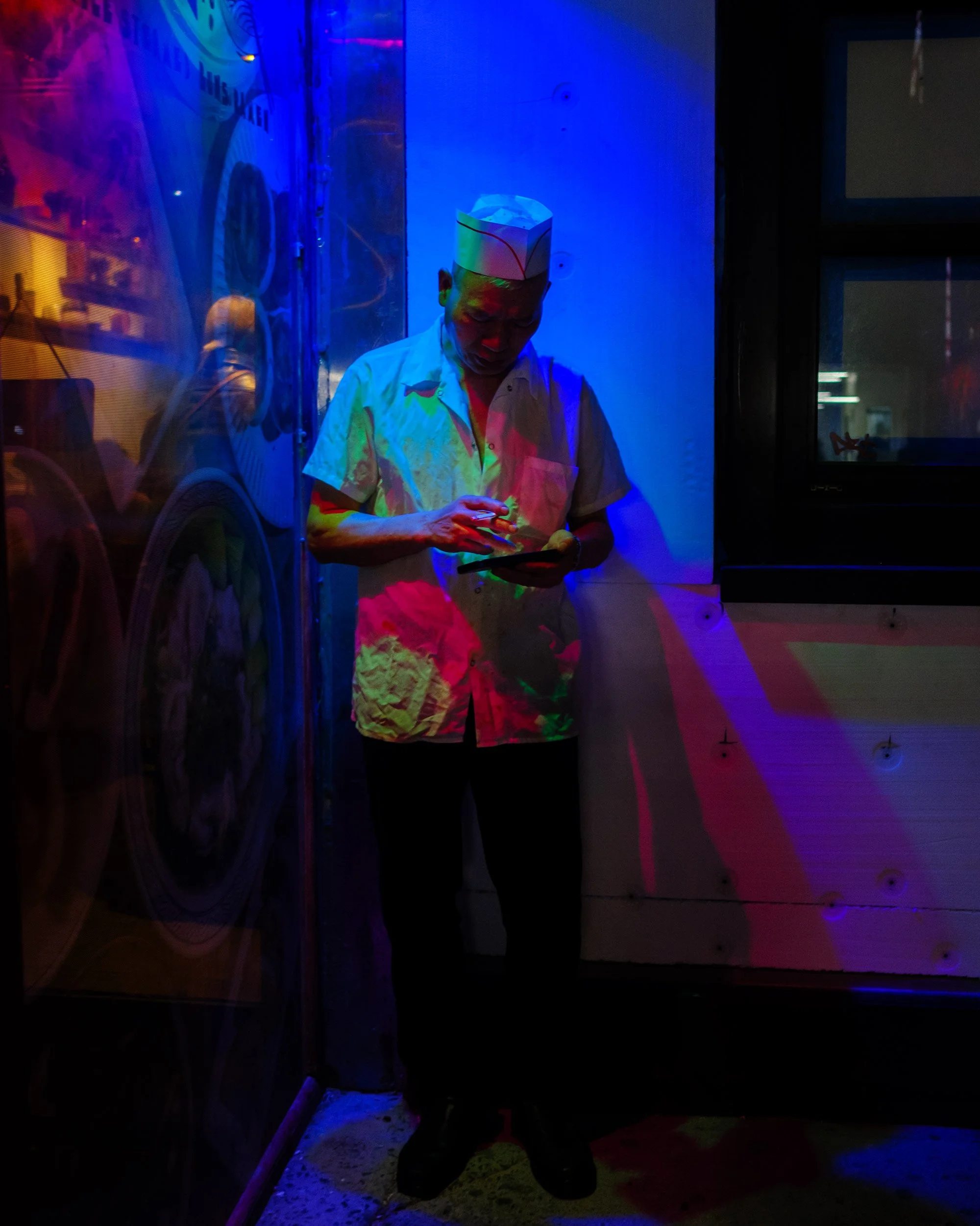 A person wearing a white chef jacket and a paper hat standing in a room with colorful neon lighting that casts various colors on their face and body, with a glass door on the right side.