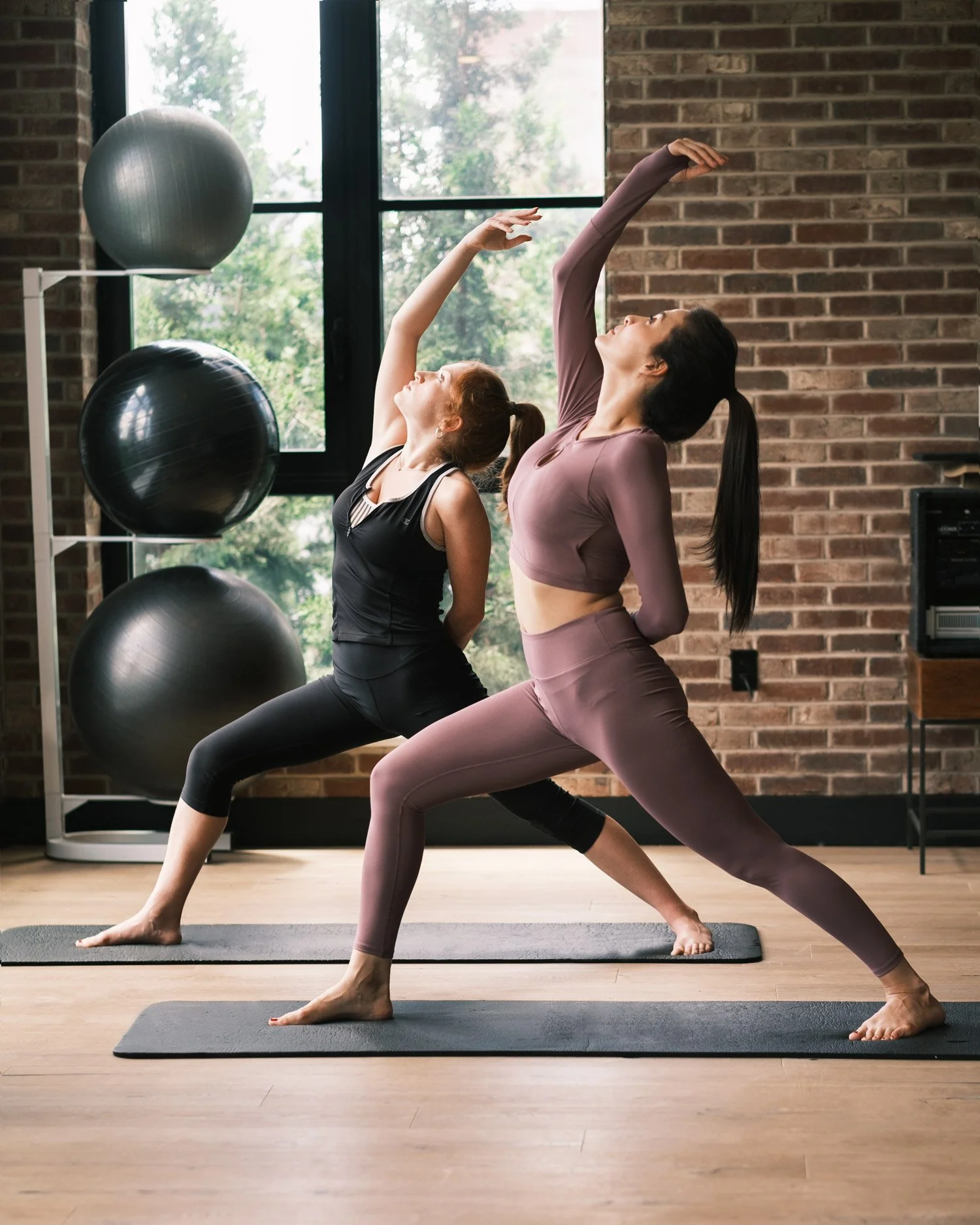 Two women practicing yoga in a studio with large windows, brick walls, and exercise balls, performing a side bend stretch on yoga mats.