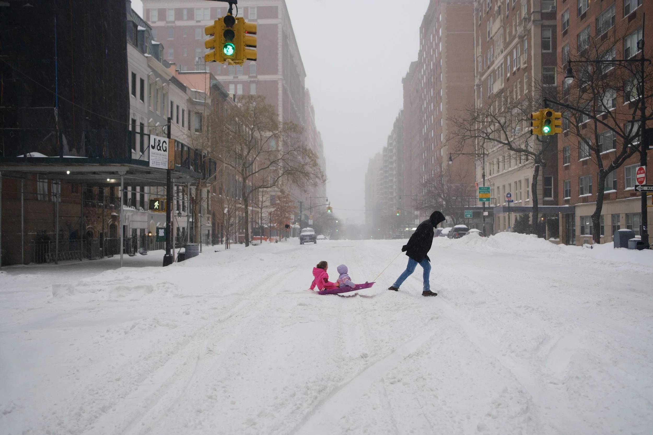 A person pulling a pink sled with two children sitting on it through snow in an urban street flanked by tall buildings, with traffic lights and bare trees visible.