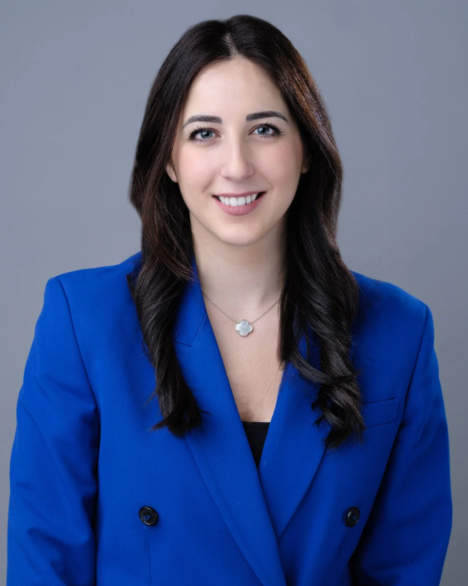 A woman with long dark hair, wearing a blue blazer, smiling, with a gray background.
