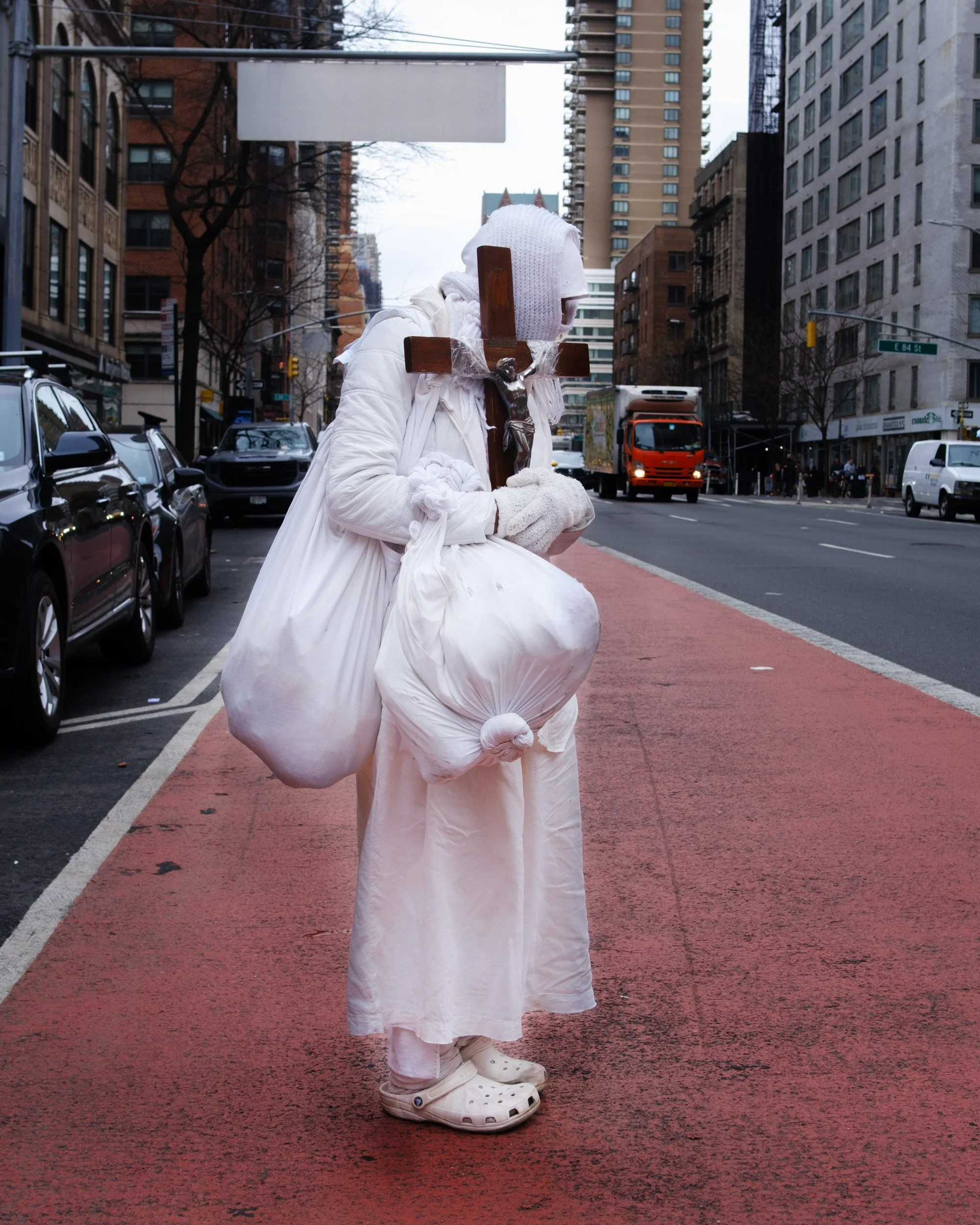 A person dressed as Jesus Christ carrying a wooden cross in an urban city street, wearing a white robe and shoes, with tall buildings and cars in the background.