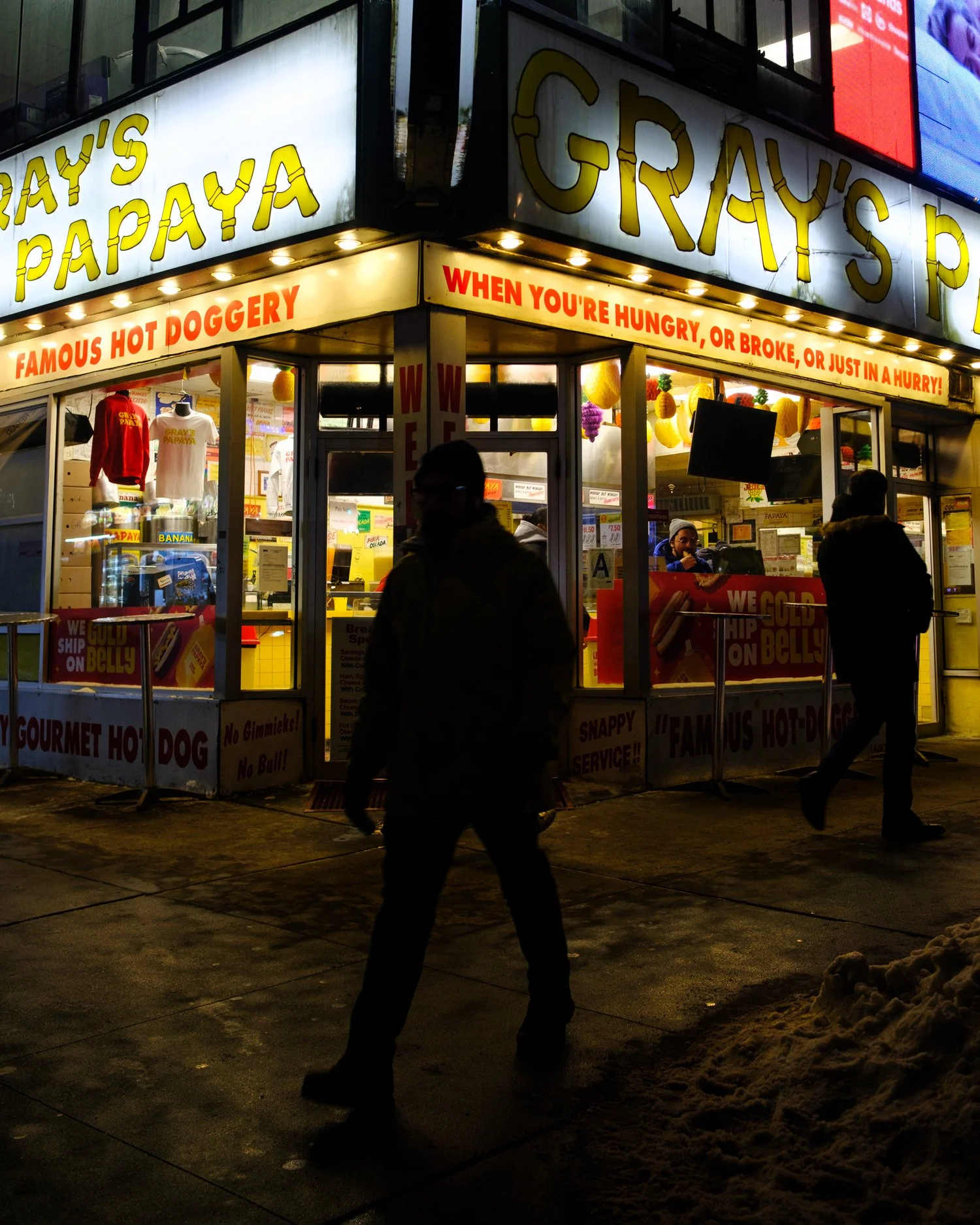 Nighttime scene of a fast-food stand named 'Gray's Papaya' with bright lights and promotional signs. Several people walk past the stand, and there is snow on the ground.