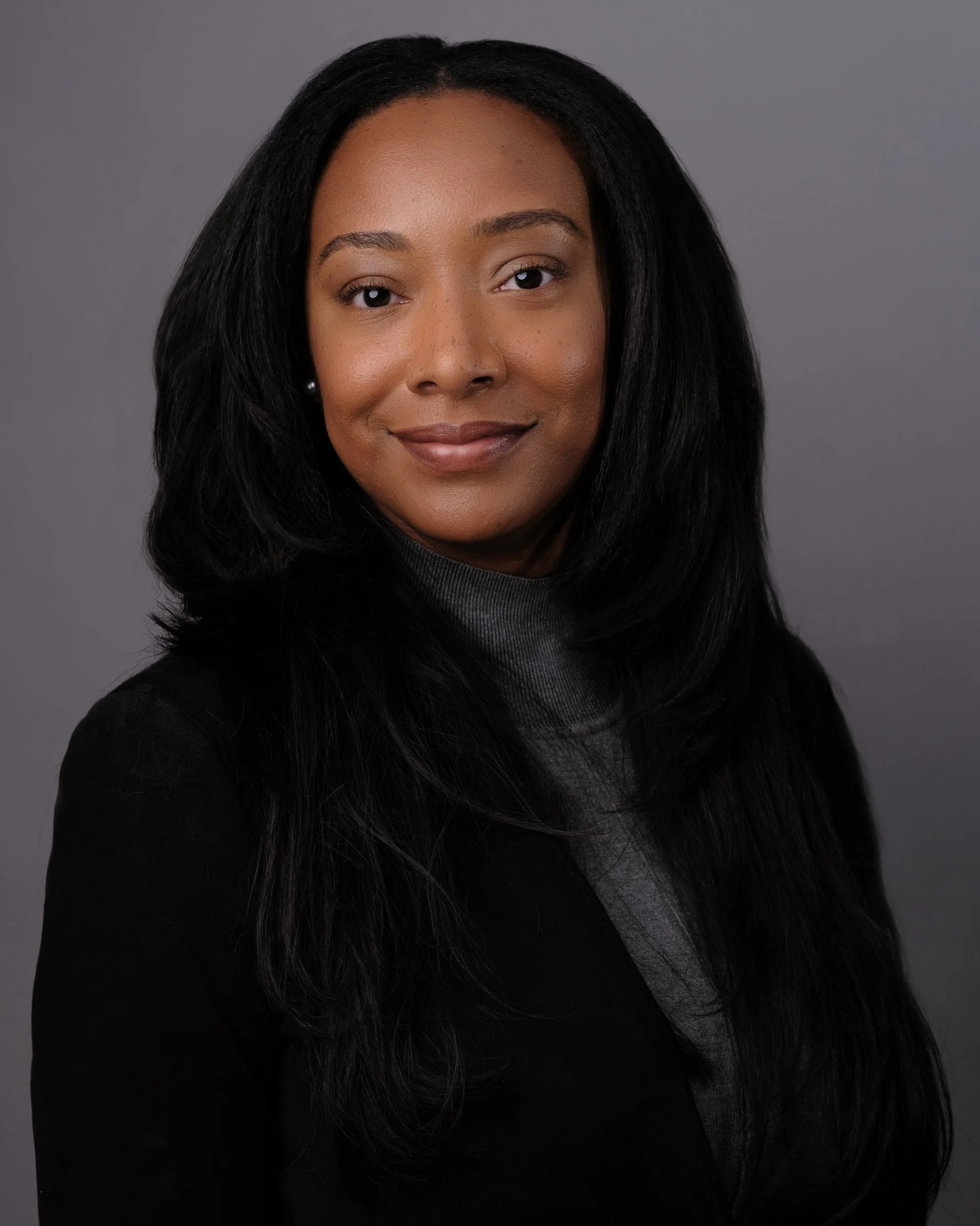 A portrait of an African American woman with long black hair, smiling, against a gray background, wearing a black top.