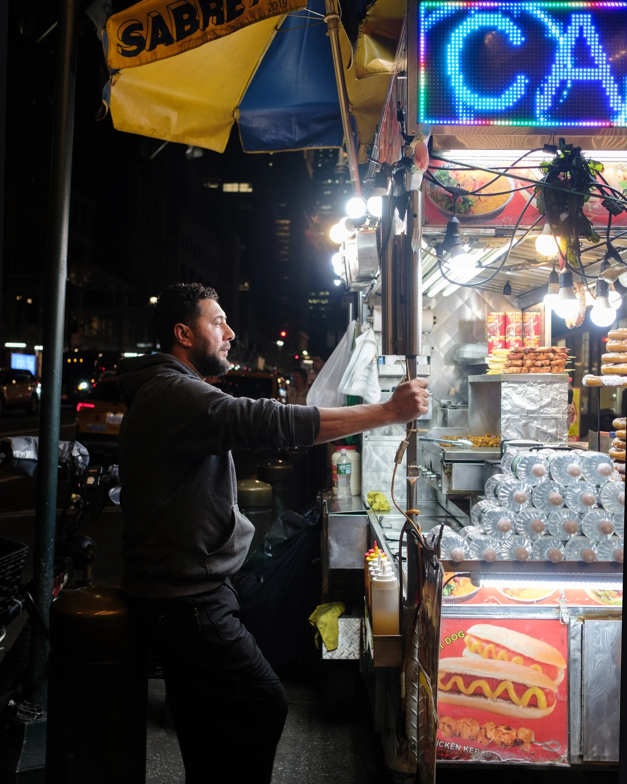 A man at a night street food stand with a yellow and blue umbrella, illuminated signs, and a display of hot dogs and fried foods.